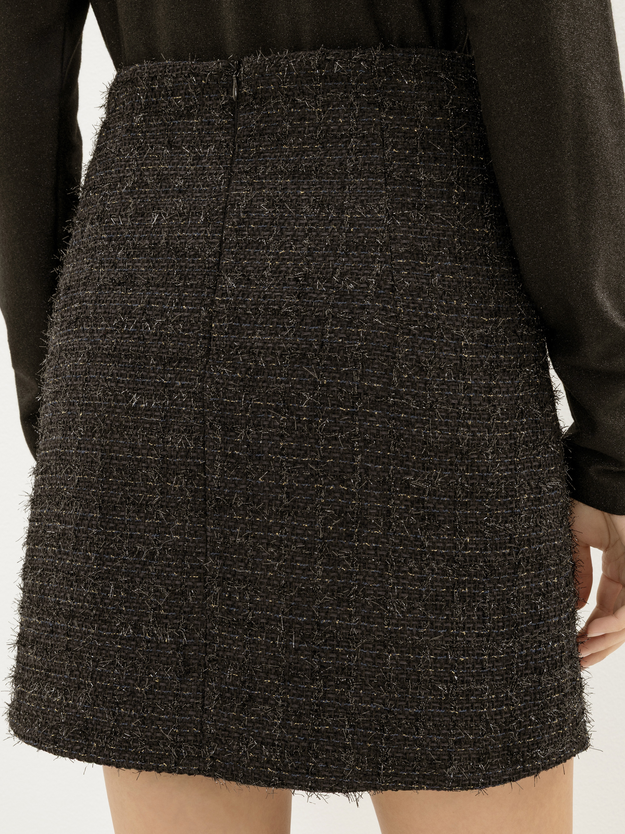 Nahaufnahme einer Person, die einen strukturierten schwarzen Tweed-Minirock und ein langärmeliges schwarzes Oberteil trägt, von der Taille abwärts und von hinten gesehen.