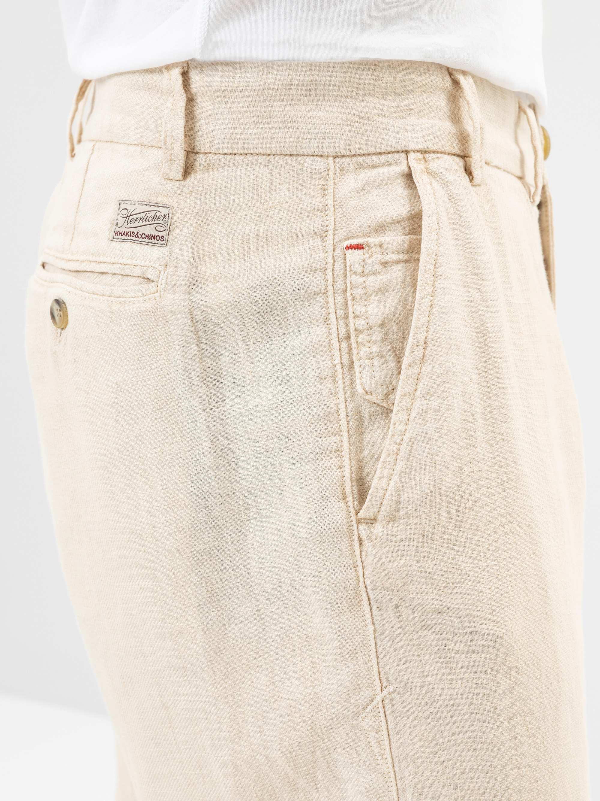 Nahaufnahme einer Person, die eine hellbeige Hose und ein weißes Hemd trägt. Zu sehen sind eine Gesäßtasche mit einem Knopf und einem kleinen Etikett darüber sowie eine Vordertasche mit sichtbaren Nähten.