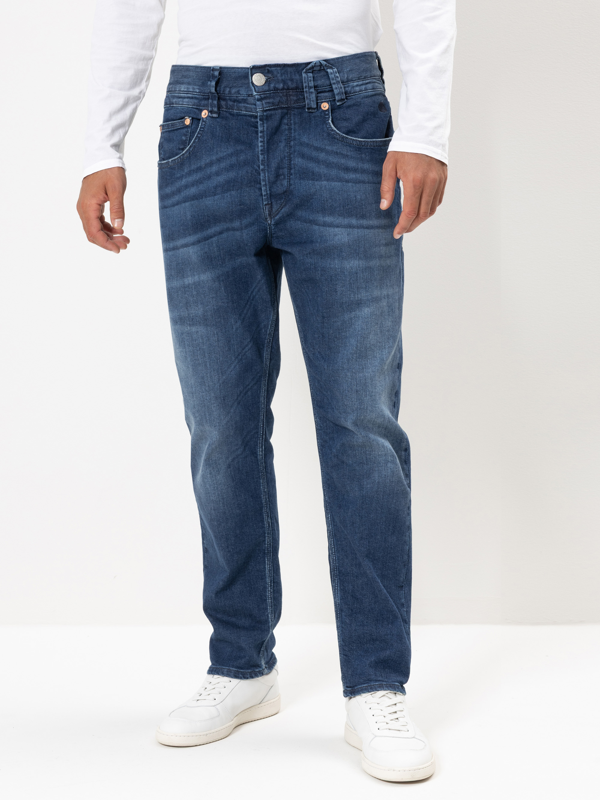 Eine Person, die eine blaue Jeans mit geradem Bein, ein weißes langärmeliges Hemd und weiße Turnschuhe trägt, steht vor einem schlichten weißen Hintergrund. Nur der Körper von den Schultern abwärts ist sichtbar.