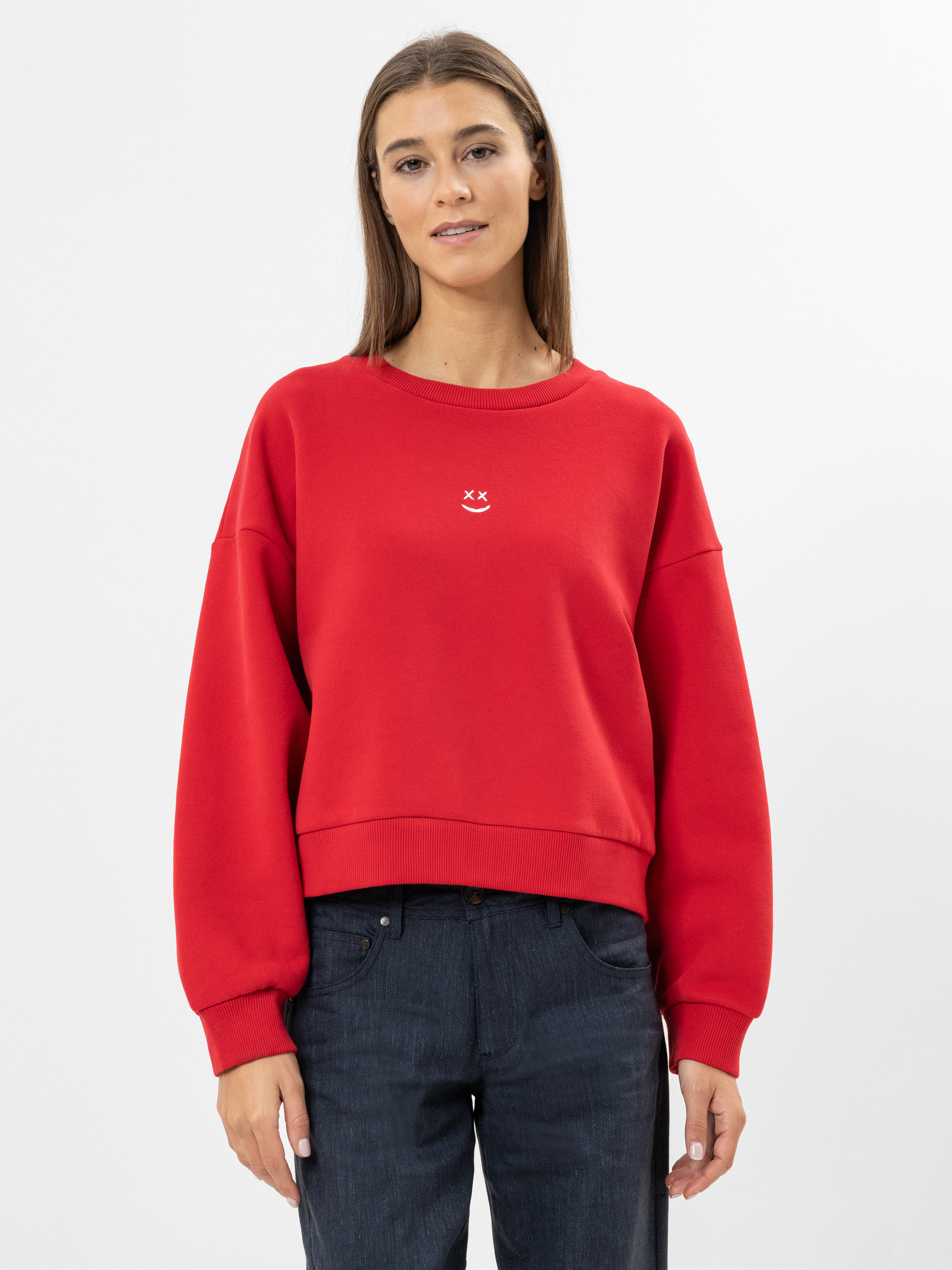 Eine Frau mit glattem braunem Haar trägt ein rotes Sweatshirt mit einem kleinen weißen Smiley-Motiv auf der Brust und dunkle Jeans vor einem schlichten weißen Hintergrund.