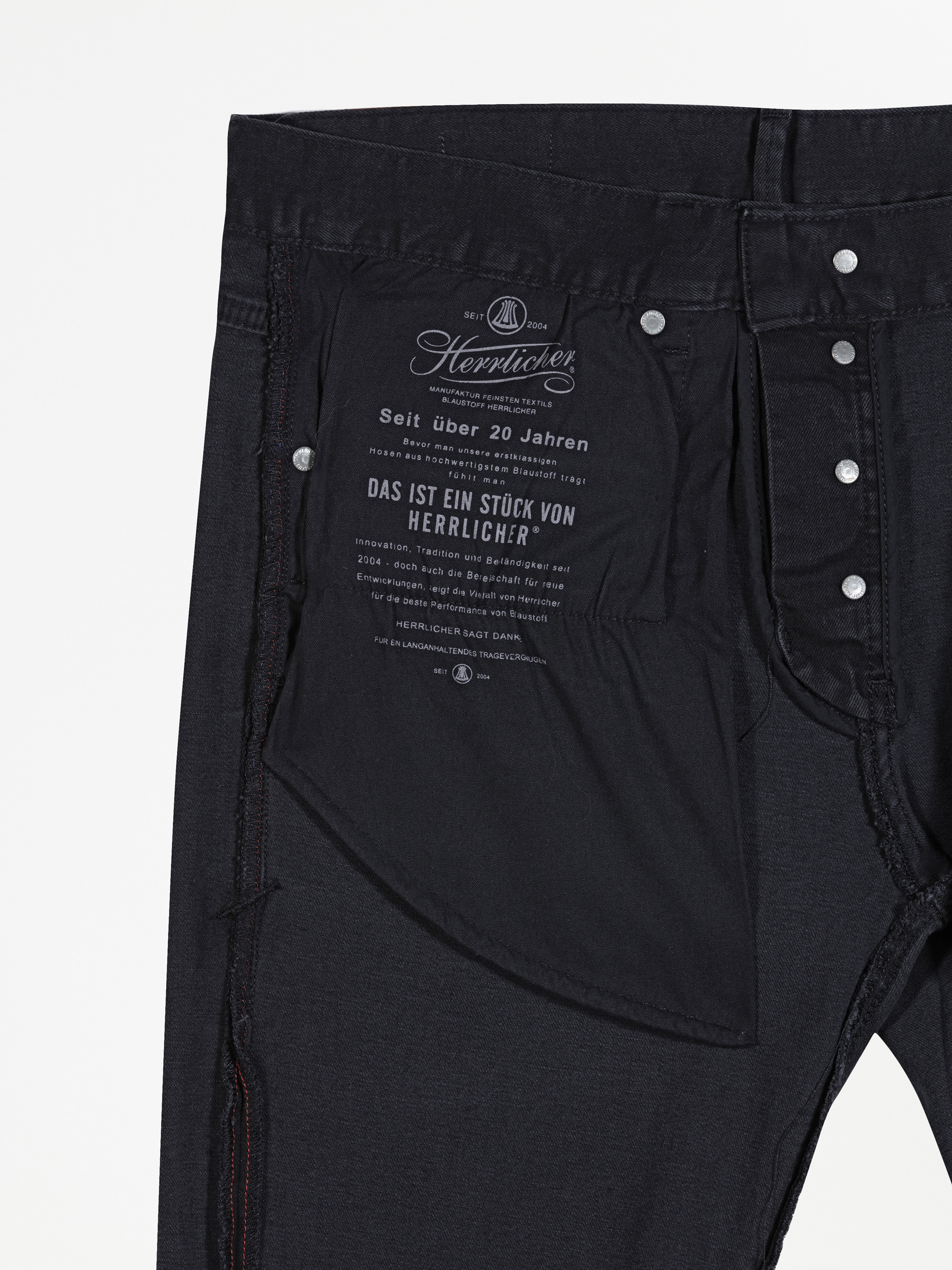Nahaufnahme der Innenseite einer schwarzen Jeans, die ein Stoffetikett mit weißem deutschem Text und silberne Nietenknöpfe am Bund und am Hosenschlitz zeigt. Das Taschenfutter ist teilweise sichtbar.