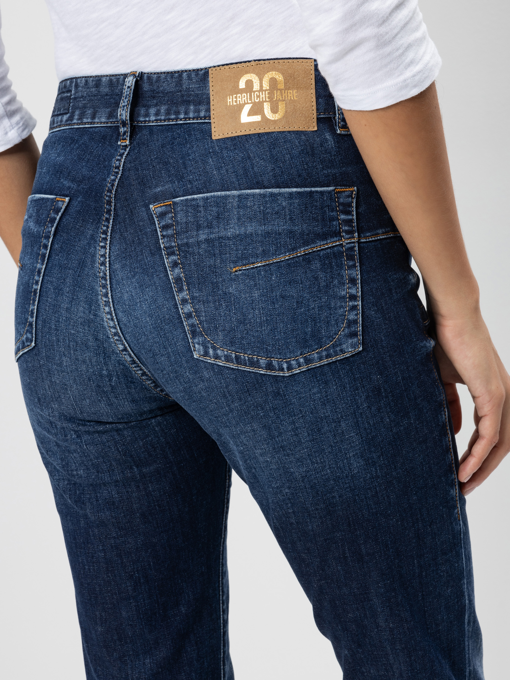 Eine Person in einer dunkelblauen Jeans und einem weißen Hemd, von hinten gesehen. Die Jeans hat einen braunen Lederaufnäher am Bund mit der Aufschrift 20 HERRLICHE JAHRE.