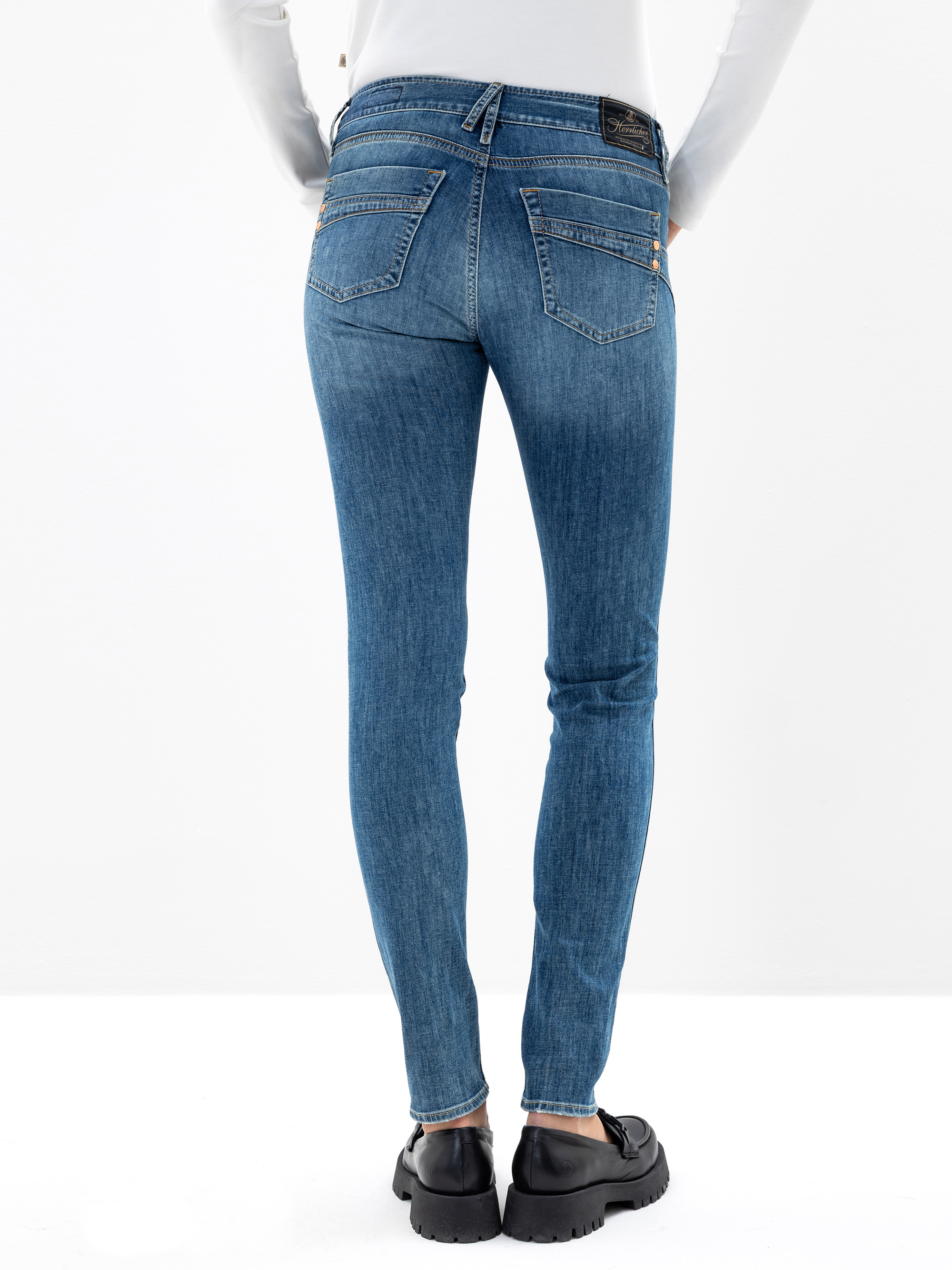 Mit dem Gesicht zur Wand stehende Person, die eine blaue Skinny-Jeans, ein weißes langärmeliges Oberteil und schwarze Schuhe trägt, dargestellt vor einem schlichten weißen Hintergrund. Die Jeans hat zwei Gesäßtaschen und sichtbare Nähte.