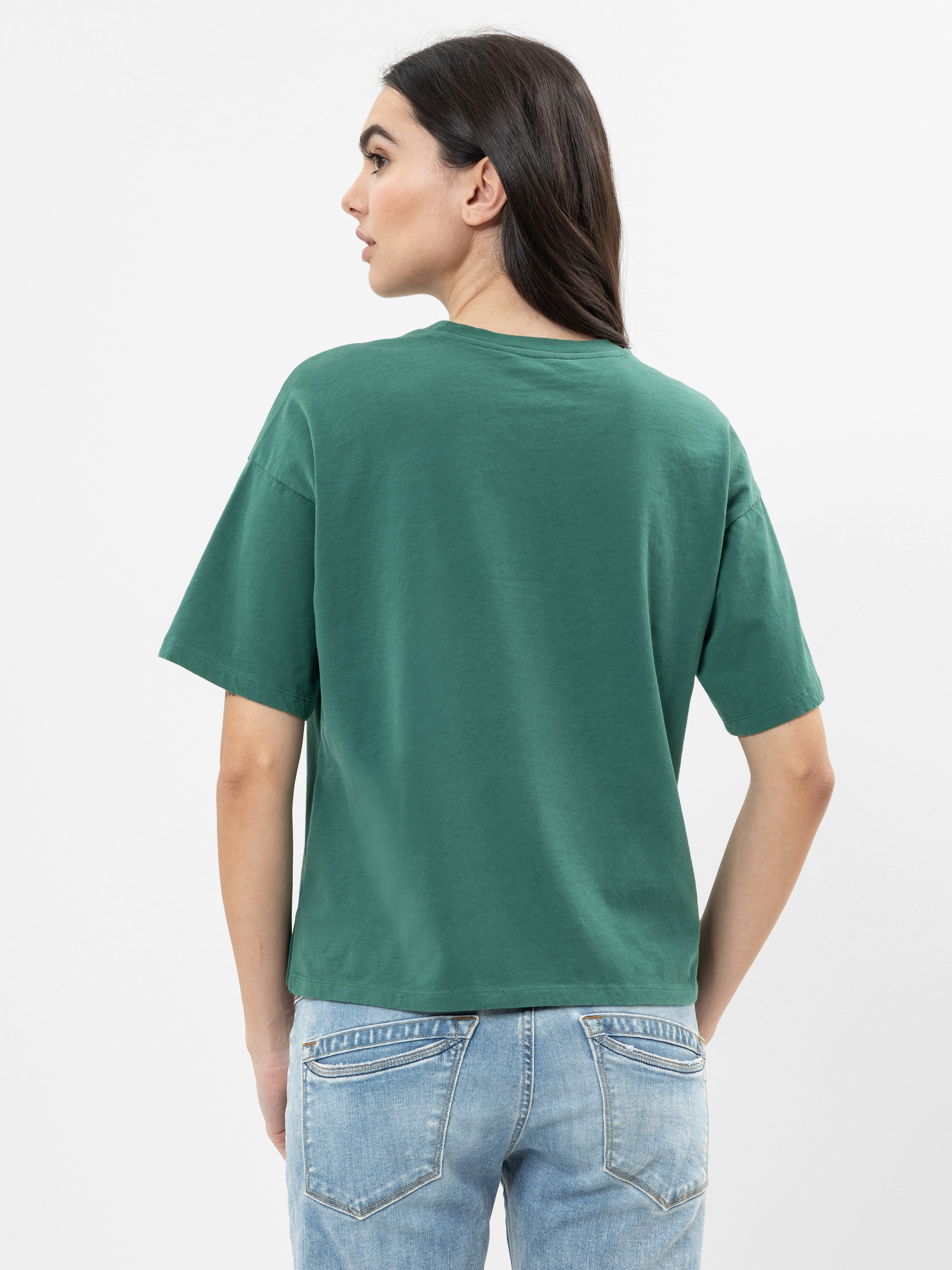 Eine Frau mit langen dunklen Haaren, die ein grünes kurzärmeliges T-Shirt und hellblaue Jeans trägt, steht mit dem Gesicht zur Kamera vor einem schlichten weißen Hintergrund.