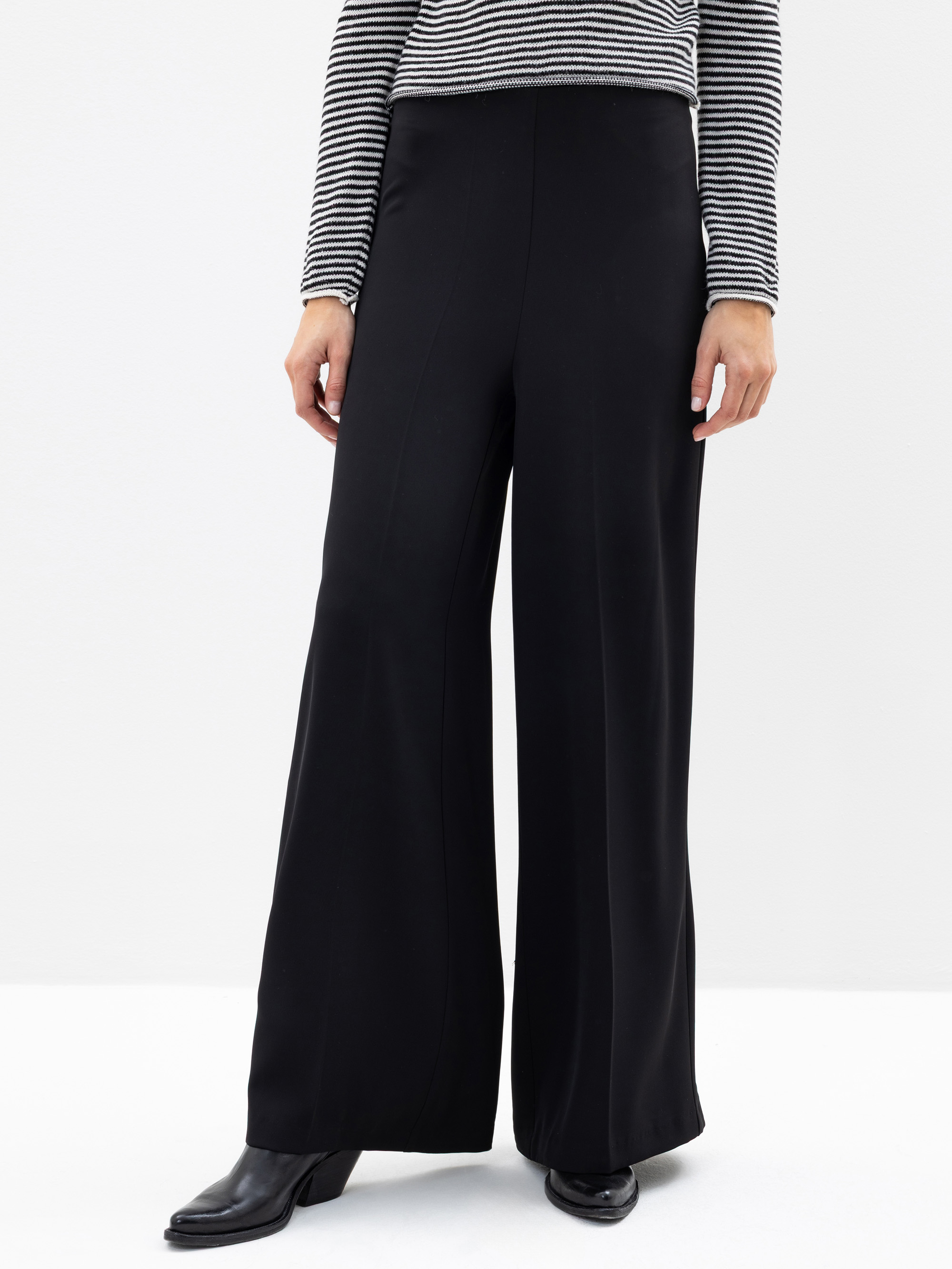 Person, die ein schwarz-weiß gestreiftes langärmeliges Oberteil, eine hochtaillierte schwarze Hose mit weitem Bein und schwarze hochhackige Stiefel trägt und vor einem schlichten weißen Hintergrund steht. Das Foto zeigt das Outfit von der Schulter bis