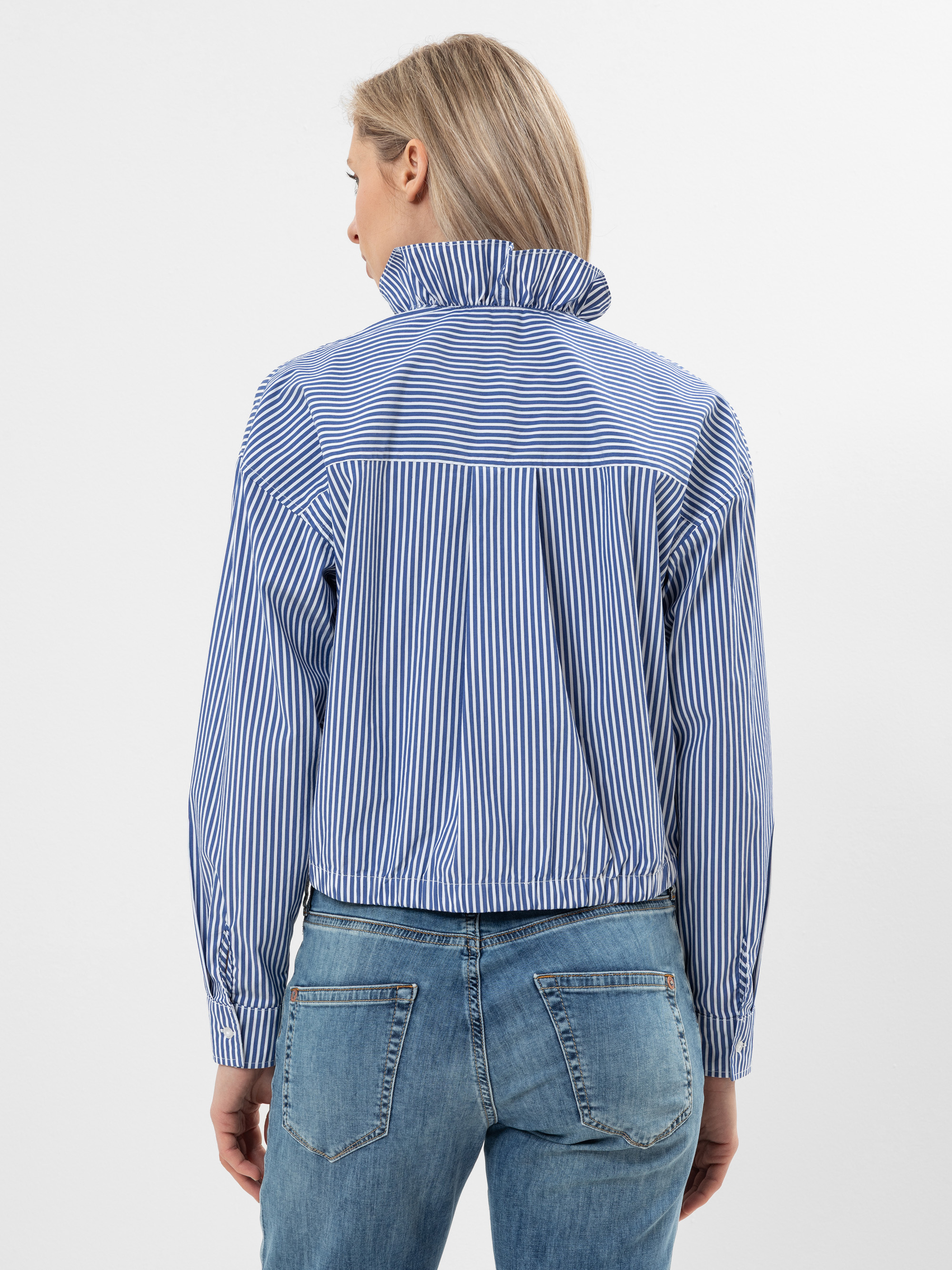 Eine Person mit langen blonden Haaren, die ein blau-weiß gestreiftes Kragenhemd und blaue Jeans trägt, steht mit dem Rücken zur Kamera vor einem schlichten weißen Hintergrund.