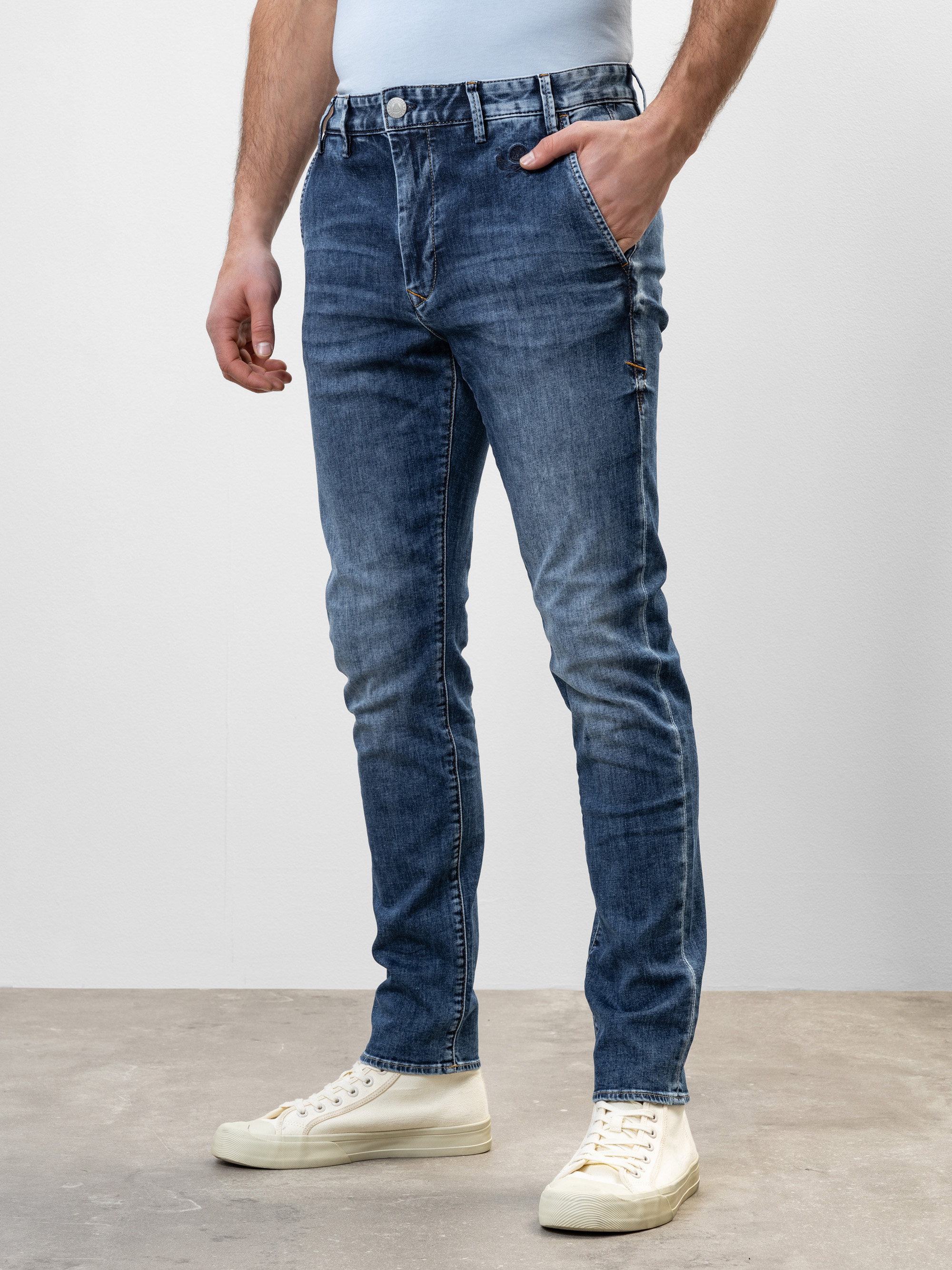 Ein Mann steht vor einem schlichten Hintergrund, trägt eine blaue Slim-Fit-Jeans, ein weißes Hemd und weiße Turnschuhe und hat die linke Hand in seiner Jeanstasche.