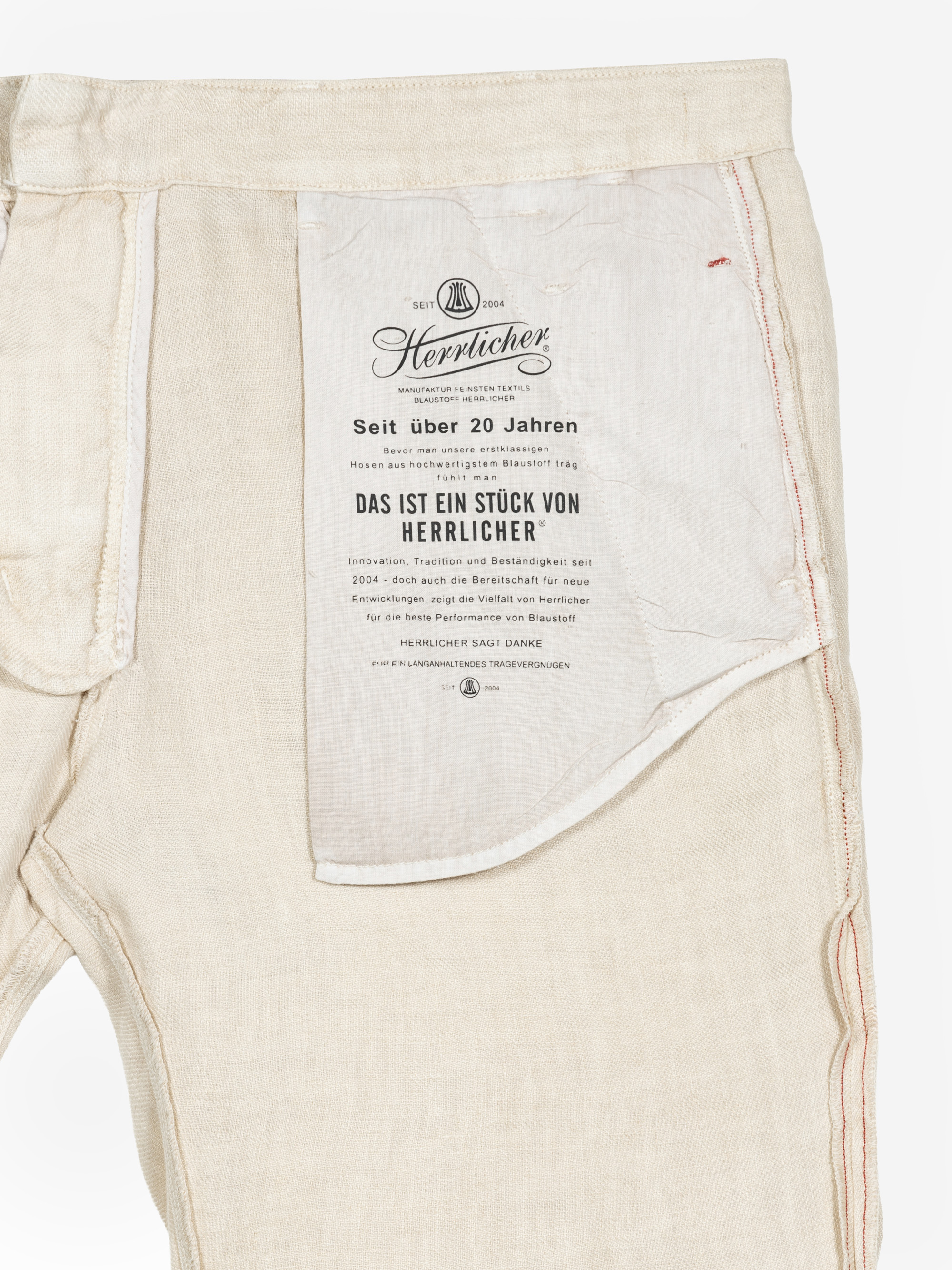 Off-White-Hose mit umgedrehter Innenseite, auf der ein großes Etikett mit deutschem Text, dem Herrlicher-Markennamen und Qualitätsangaben in schwarzer Schrift zu sehen ist. Auf der rechten Seite ist eine Naht mit roten Nähten zu sehen.