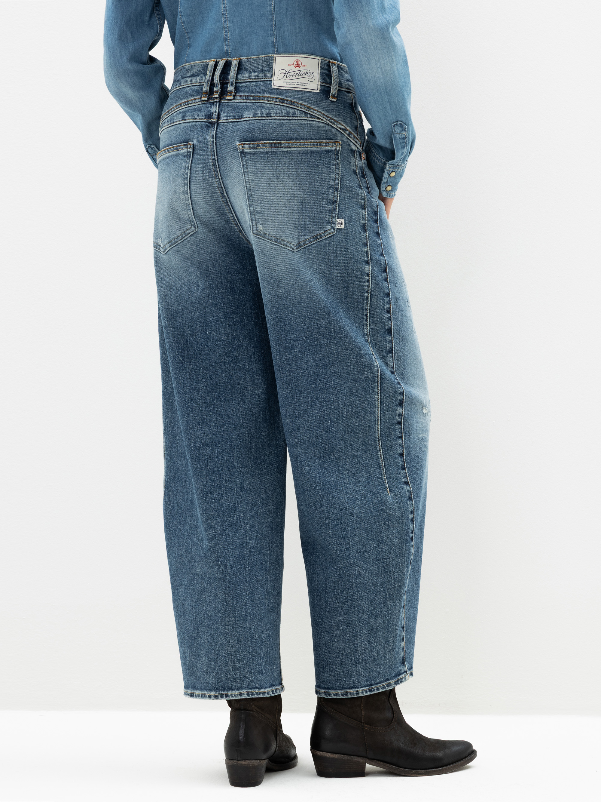 Eine Person, die locker sitzende, hochtaillierte Bluejeans, ein passendes Jeanshemd und schwarze Stiefeletten trägt, wird von hinten vor einem schlichten weißen Hintergrund gezeigt.