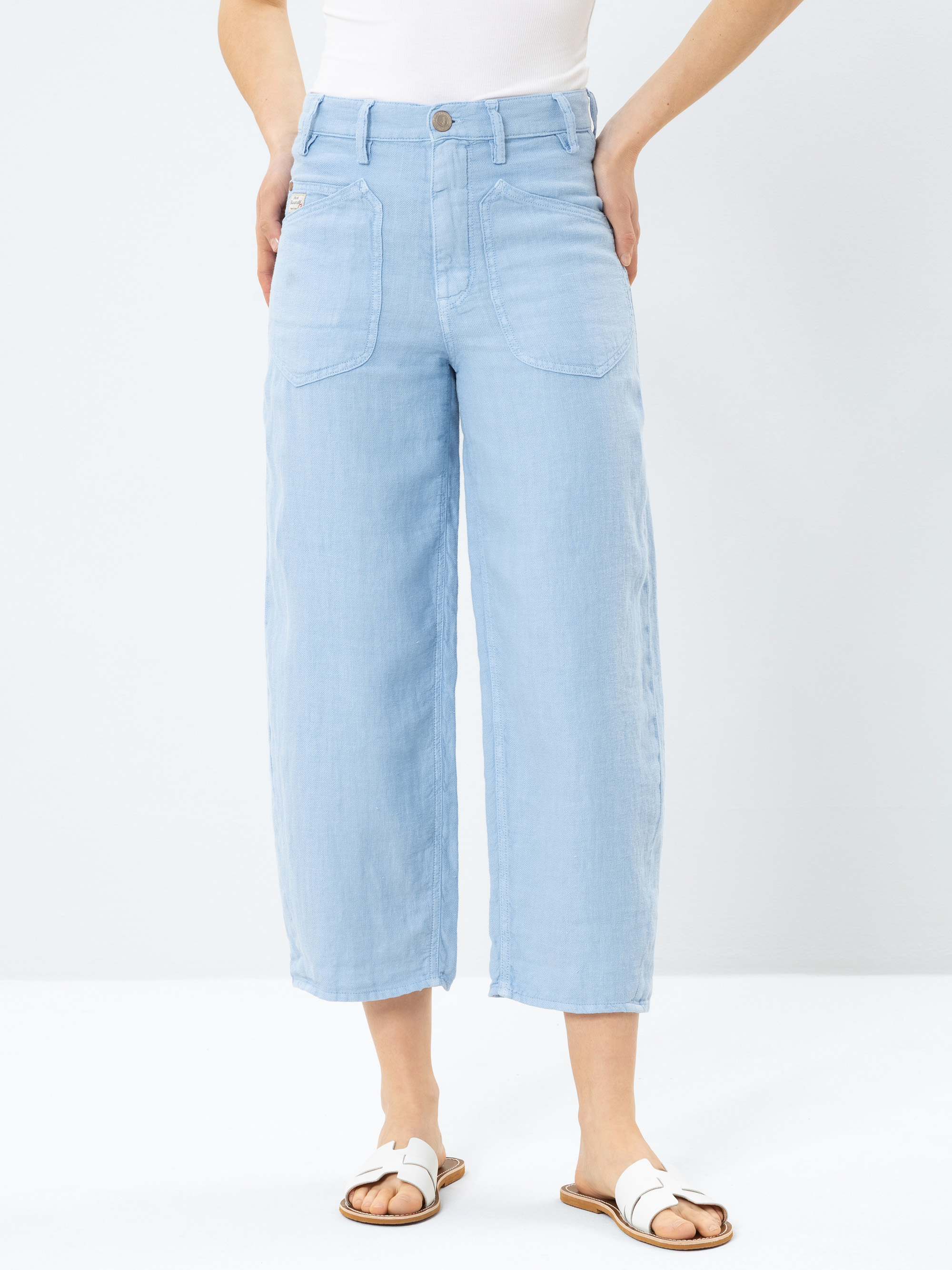 Person, die hellblaue Jeans mit weitem Bein und großen Vordertaschen, ein weißes ärmelloses Oberteil und weiße Sandalen trägt und vor einem schlichten weißen Hintergrund steht. Die Hände sind in die Hüften gestemmt.