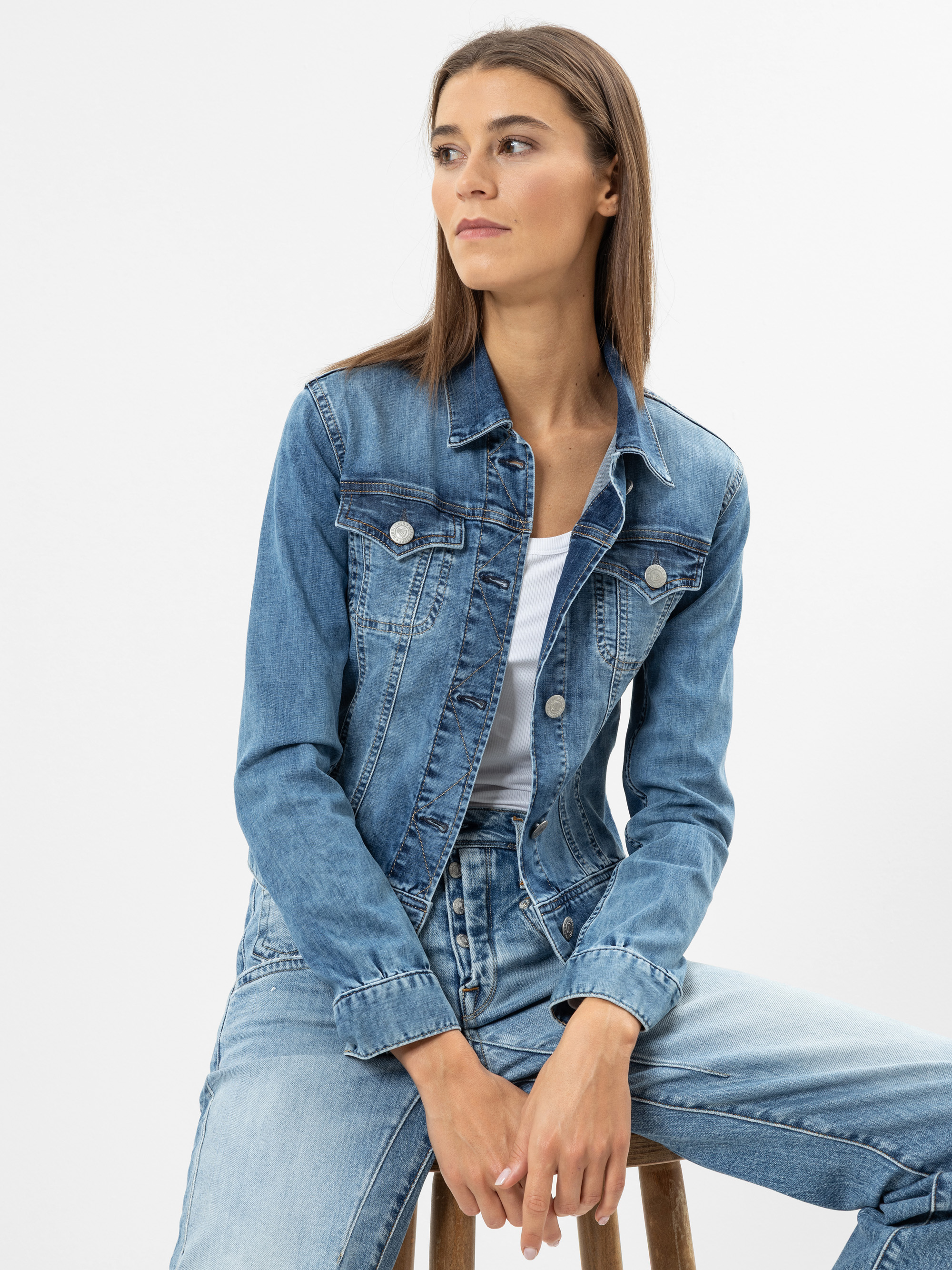 Eine Frau mit glattem braunem Haar, die eine blaue Jeansjacke über einem weißen Oberteil und blaue Jeans trägt, sitzt auf einem Hocker und blickt vor einem schlichten hellen Hintergrund zur Seite.