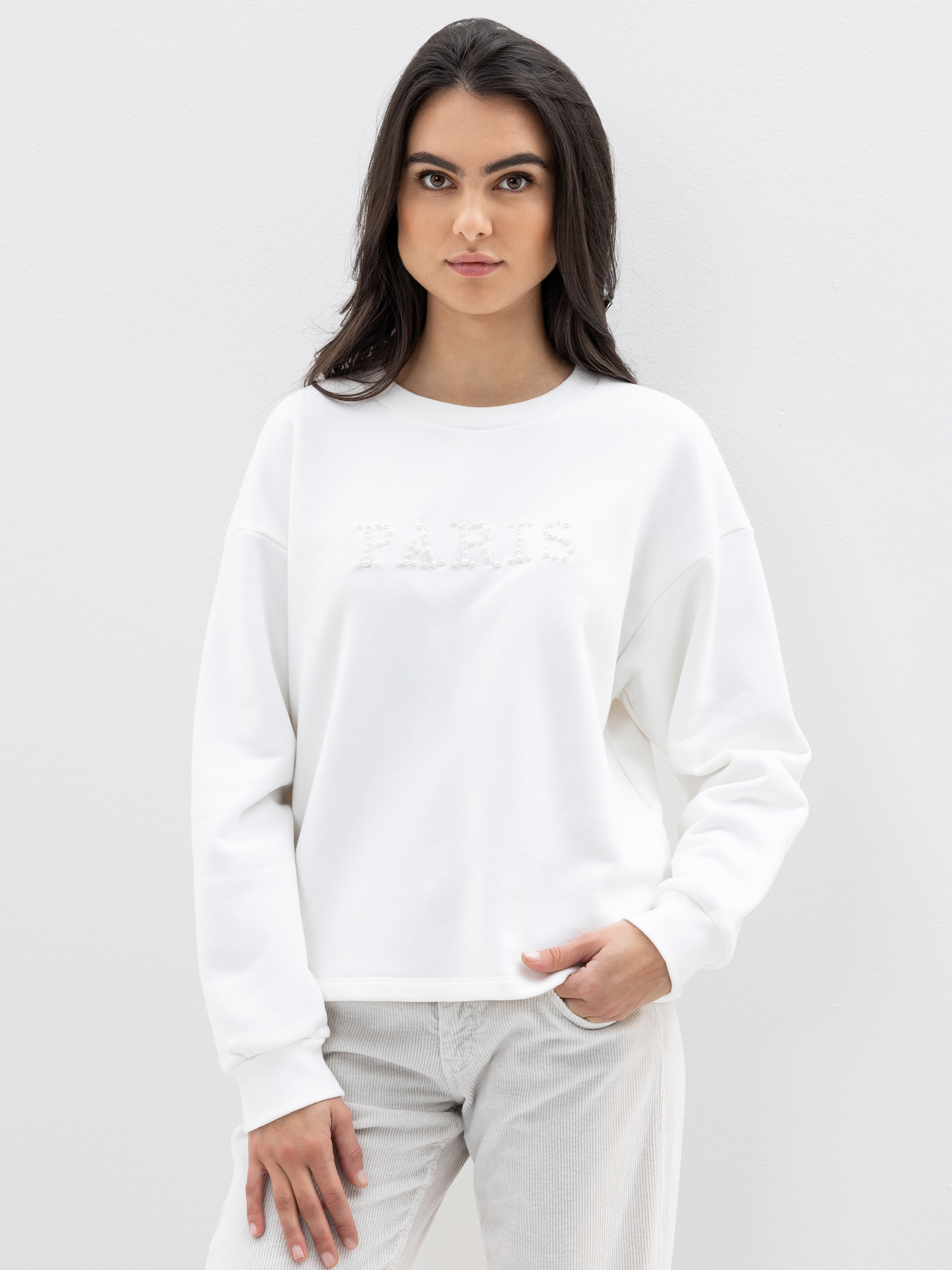 Eine Frau mit langen dunklen Haaren trägt ein weißes Sweatshirt mit der Aufschrift PARIS in strukturierten Buchstaben und eine helle Hose. Sie steht vor einem schlichten weißen Hintergrund und blickt mit neutralem Blick direkt in die Kamera.
