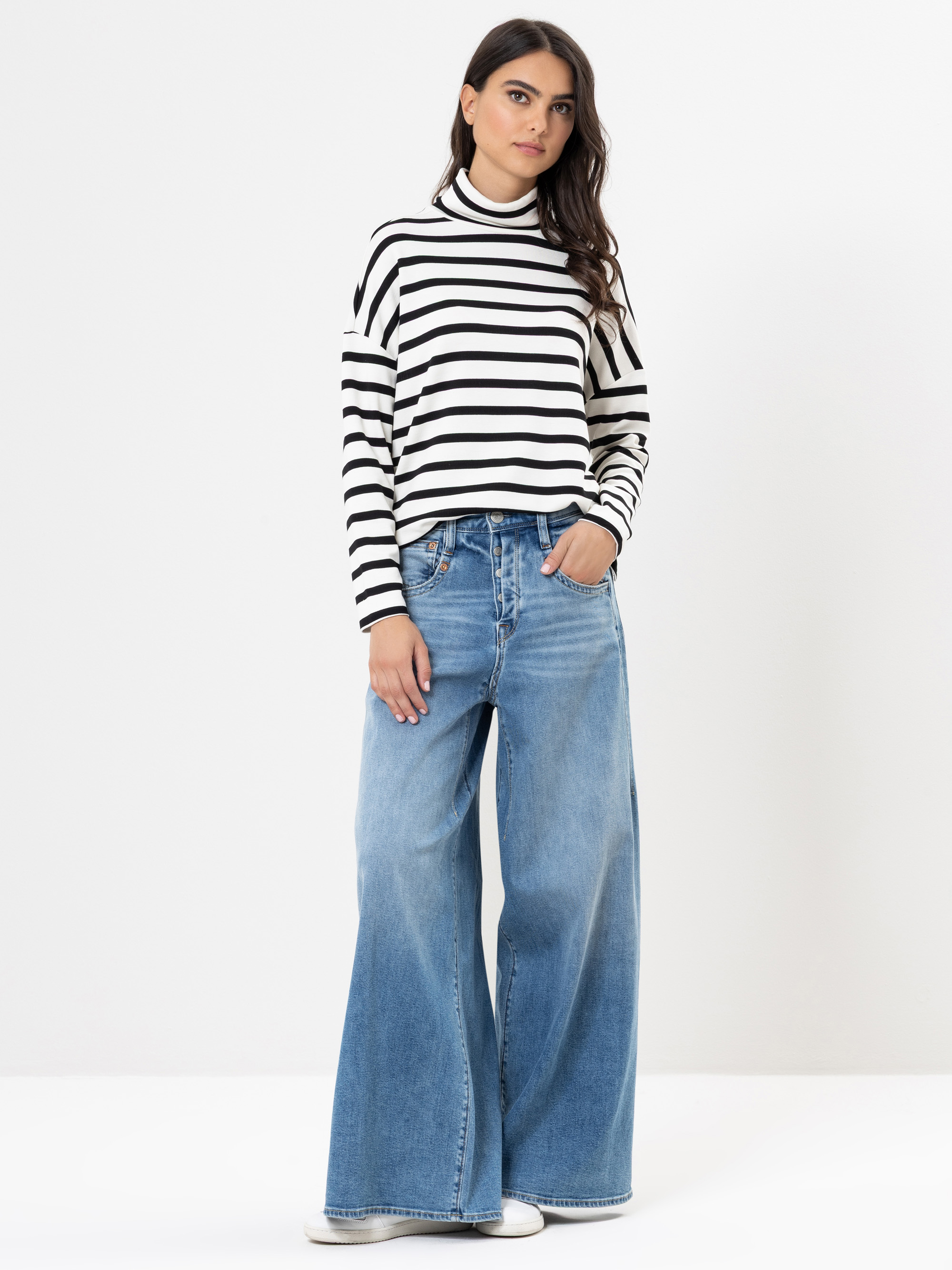 Eine Frau steht vor einem schlichten weißen Hintergrund und trägt einen schwarz-weiß gestreiften Rollkragenpullover und eine weit geschnittene Bluejeans. Sie hat langes, dunkles Haar und weiße Turnschuhe. Sie hat die Hände in den Taschen und schaut i