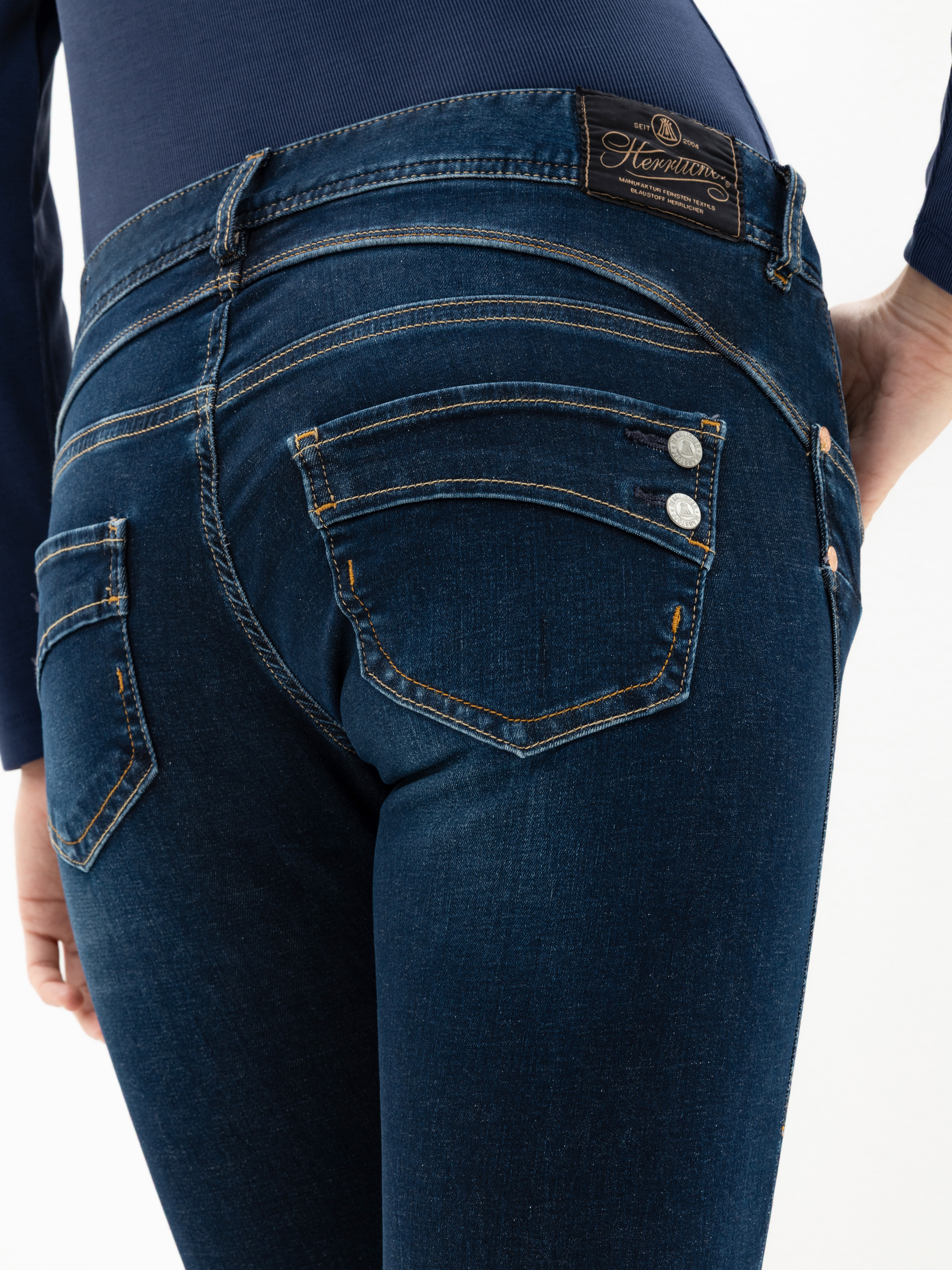 Nahaufnahme einer Person, die eine dunkelblaue Denim-Jeans mit Ziernähten, Metallnieten und einem braunen Hermans-Markenetikett am Bund trägt. Die Hand der Person ruht auf ihrer Hüfte.