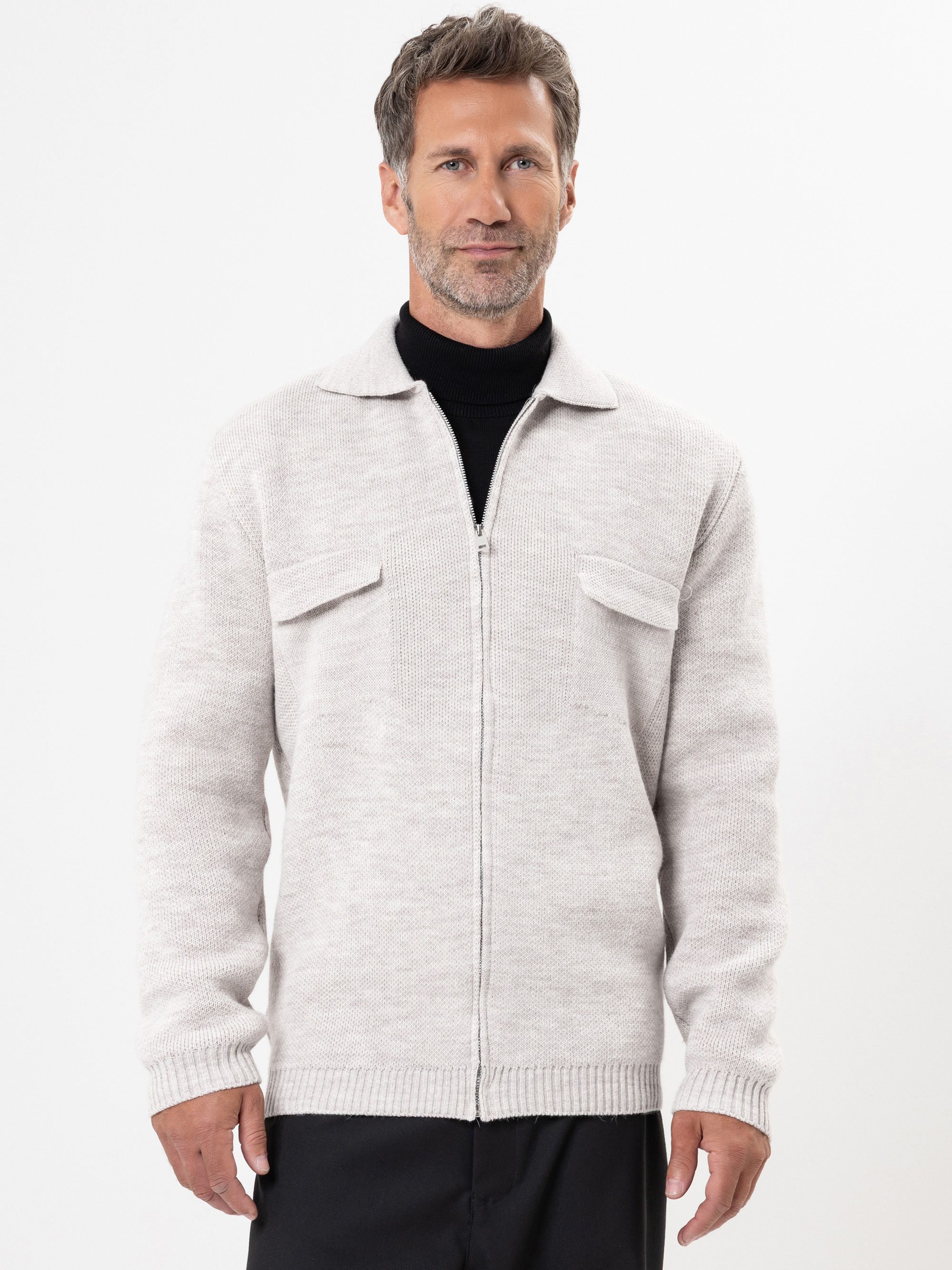 Ein Mann mittleren Alters mit kurzen braunen Haaren und einem Bart trägt einen hellgrauen Pullover mit Reißverschluss und zwei Brusttaschen über einem schwarzen Rollkragenpullover. Er steht mit dem Gesicht nach vorne vor einem schlichten weißen Hinter