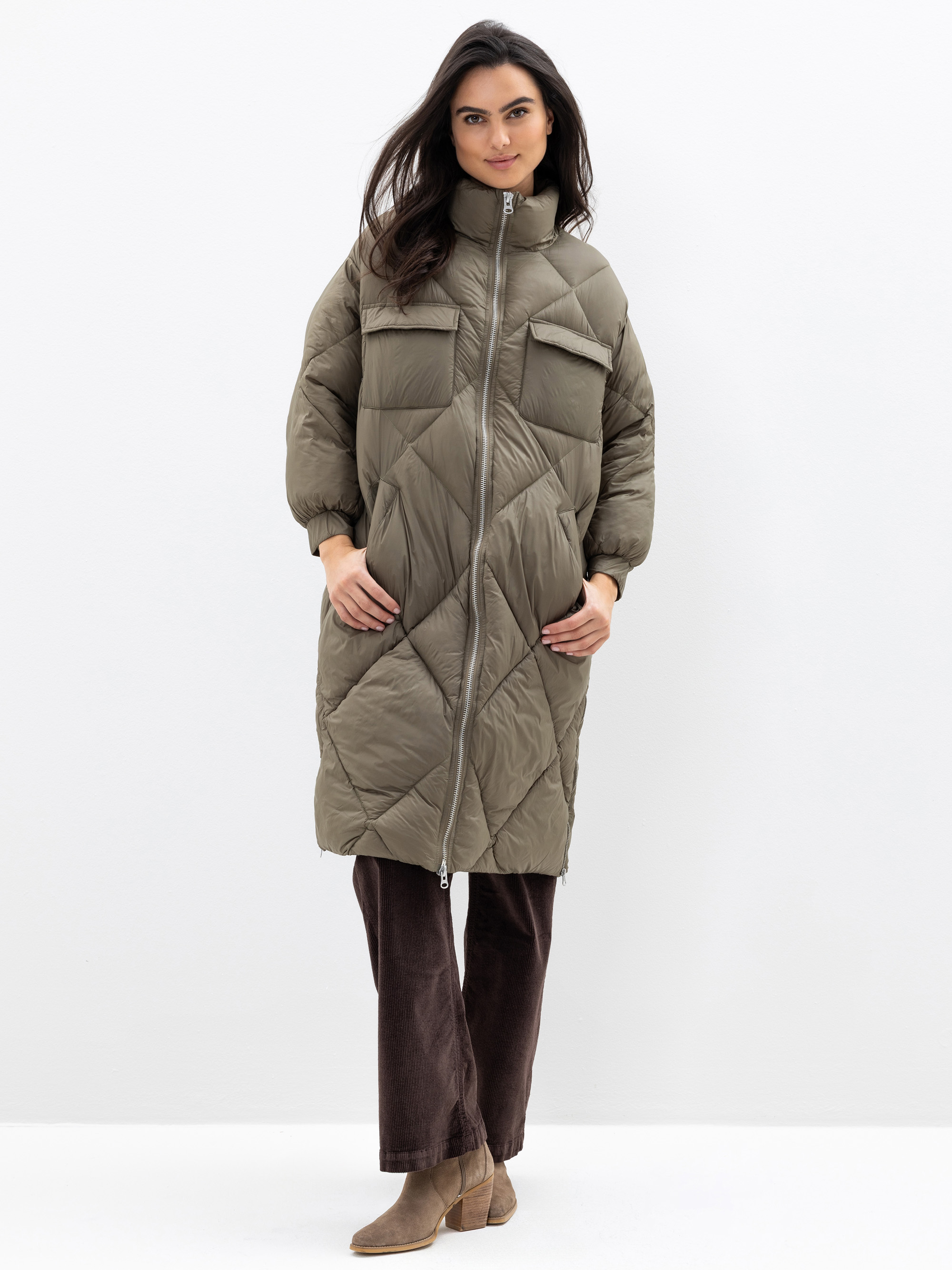 Eine Frau steht vor einem weißen Hintergrund und trägt einen langen, olivgrünen Steppmantel mit hohem Kragen, Frontreißverschluss, zwei Brusttaschen, eine braune Hose und hellbraune Stiefeletten. Sie lächelt und hat die Hände in den Manteltaschen.