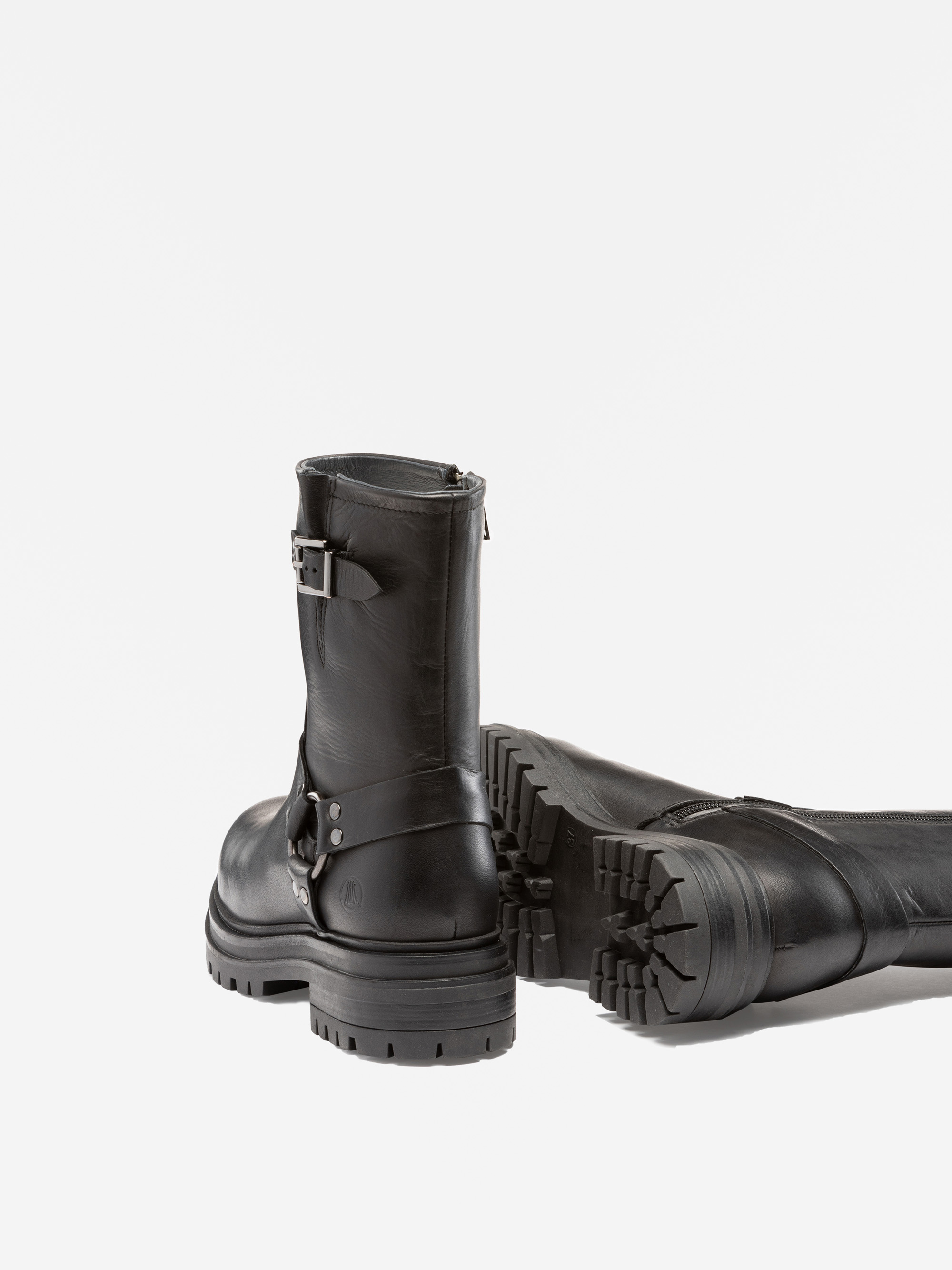 Ein Paar schwarze, halbhohe Lederstiefel mit dicken, robusten Sohlen und Metallschnallendetails, wobei ein Stiefel aufrecht steht und der andere auf der Seite liegt, beide auf weißem Hintergrund.