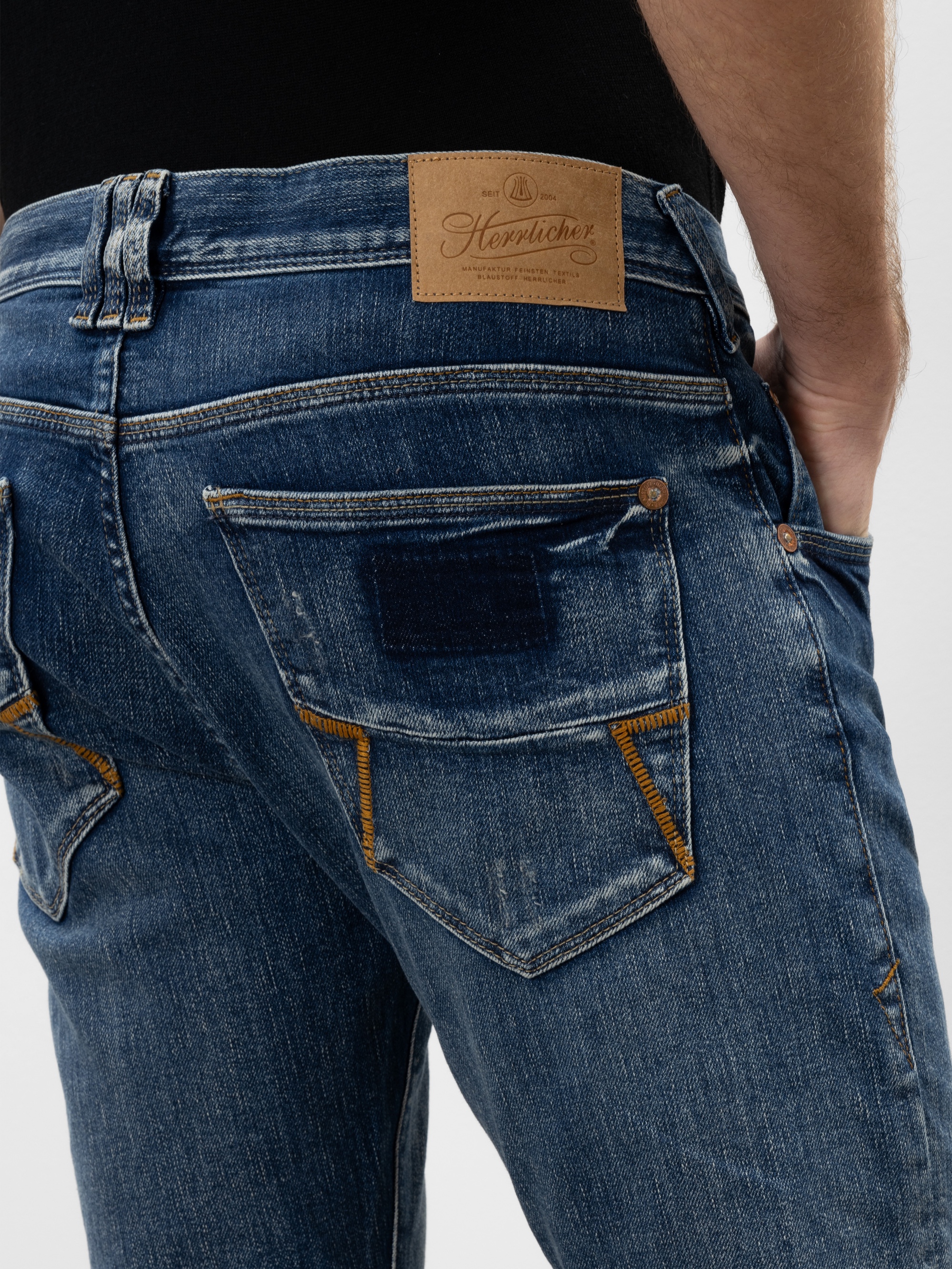 Nahaufnahme einer Person, die eine blaue Jeans mit markanten gelben Nähten und einem braunen Lederaufnäher am Bund trägt. Die Person hat eine Hand in der Hosentasche, und die Jeans wirkt leicht abgenutzt.