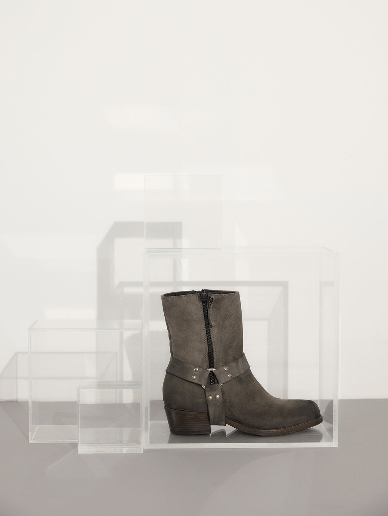Eine graue Stiefelette mit schwarzem Reißverschluss und metallischen Nieten ist in einer durchsichtigen Acrylbox ausgestellt, mit anderen leeren durchsichtigen Boxen im Hintergrund vor einem neutralen Hintergrund.