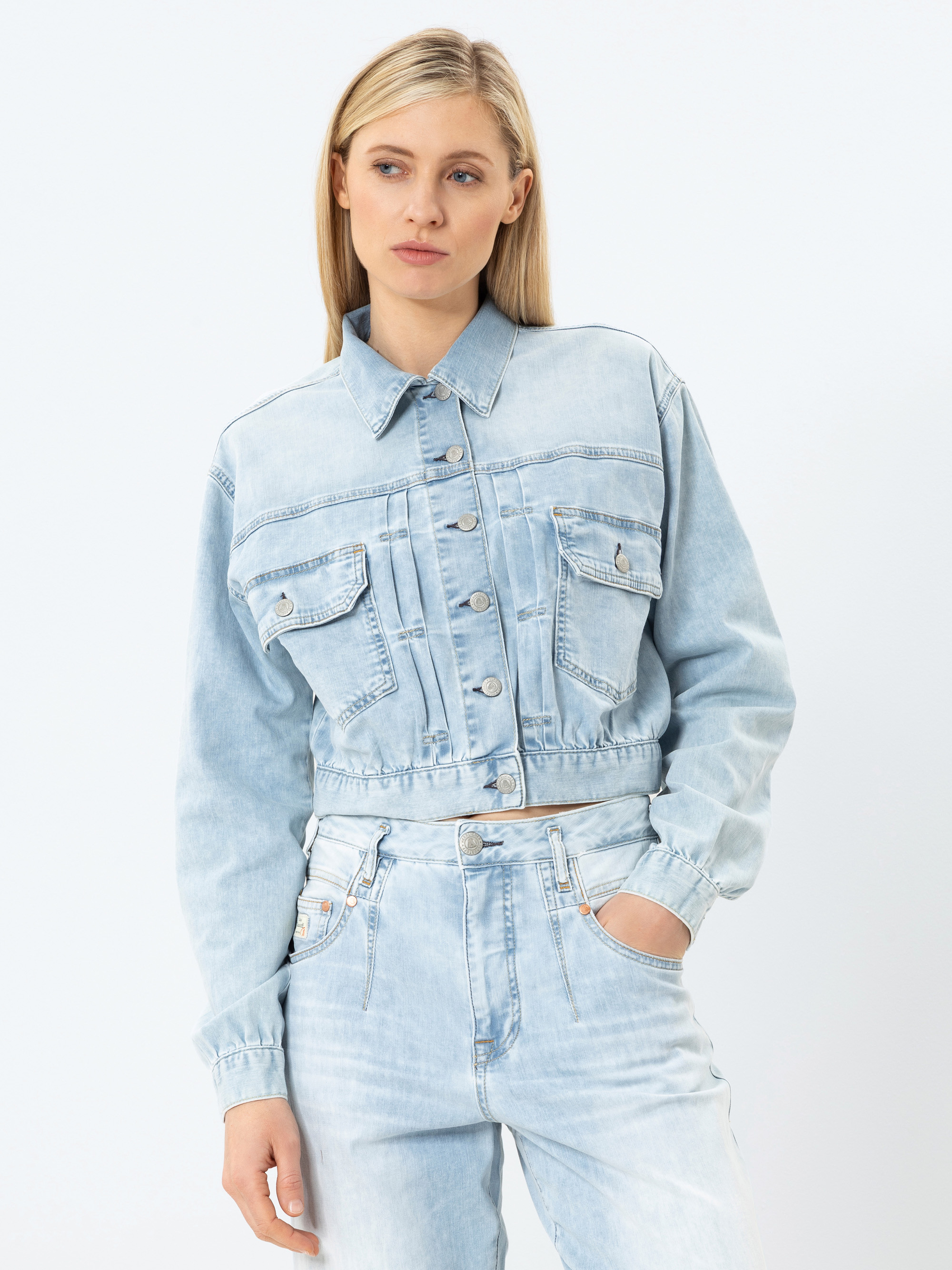 Eine Frau, die eine hellblaue Jeansjacke und eine dazu passende High-Waisted-Jeans trägt, steht vor einem schlichten weißen Hintergrund und blickt mit einem neutralen Ausdruck leicht zur Seite.