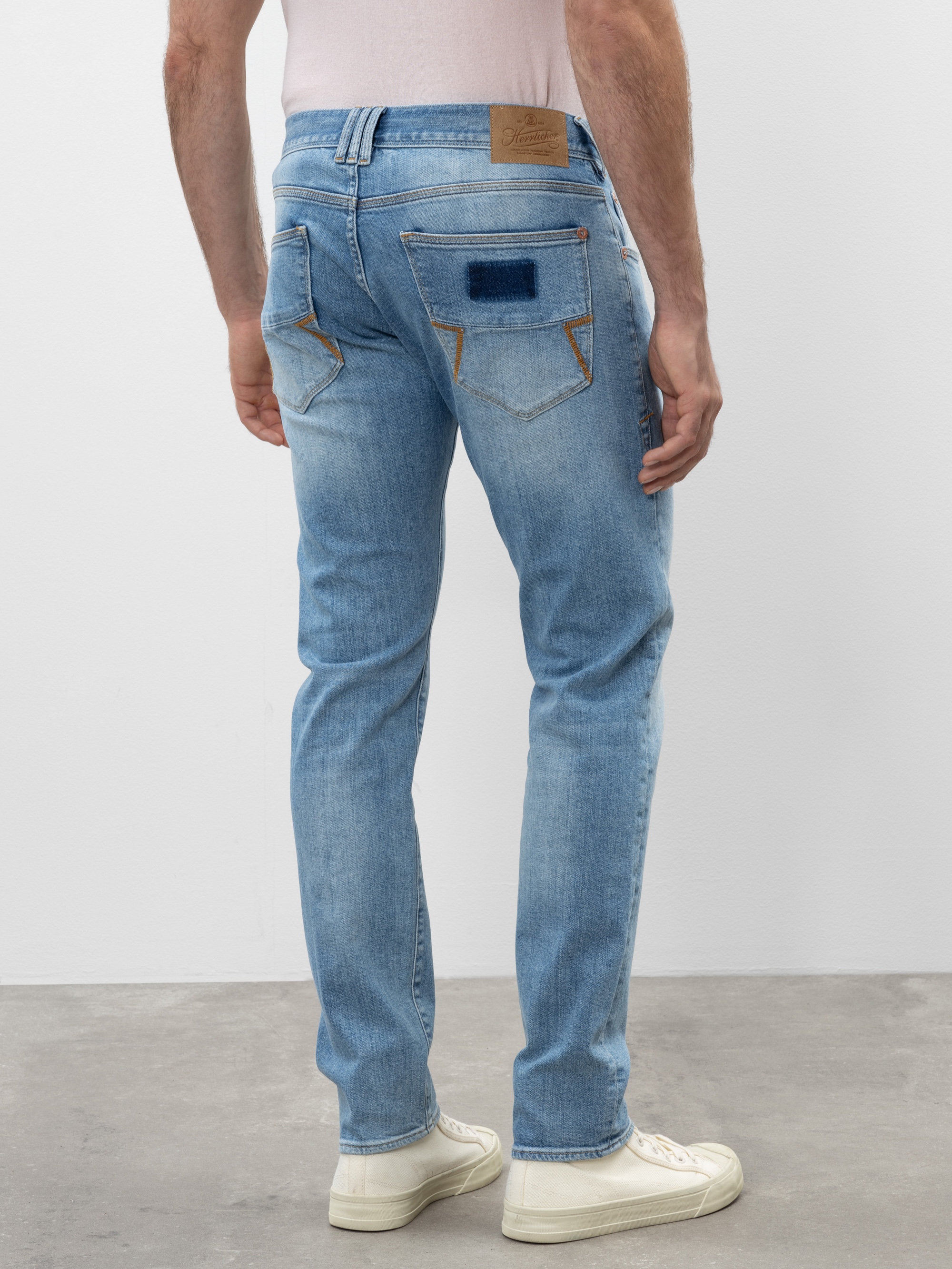 Eine Person in hellblauen Jeans mit Ziernähten und aufgesetzten Details an den Gesäßtaschen, kombiniert mit weißen Turnschuhen, steht auf einem grauen Boden vor einer schlichten weißen Wand.