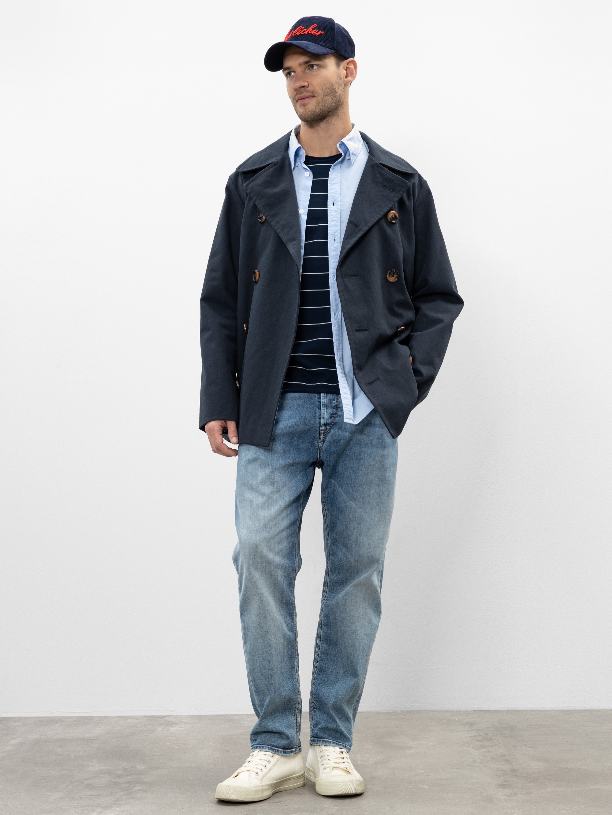Ein Mann steht vor einem schlichten Hintergrund und trägt eine schwarze Mütze, eine dunkle, marineblaue Jacke, ein hellblaues Hemd, einen marineblauen gestreiften Pullover, verwaschene blaue Jeans und weiße Turnschuhe. Eine Hand steckt in seiner Tasche