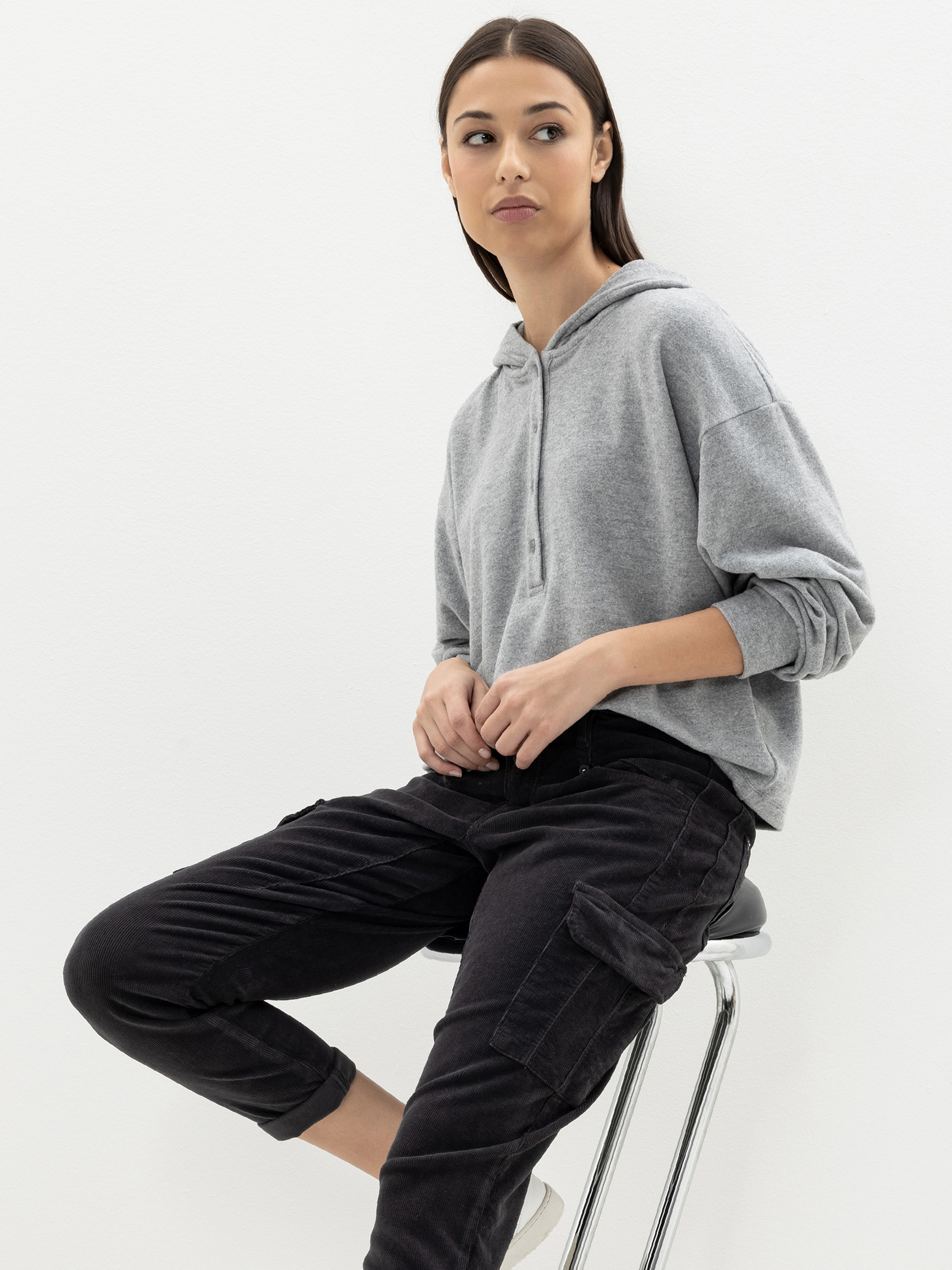 Eine Frau mit glattem braunem Haar sitzt auf einem Hocker vor einem schlichten weißen Hintergrund und trägt ein graues Sweatshirt mit Kapuze und eine schwarze Cargohose. Sie blickt leicht zur Seite und hat einen neutralen Blick.