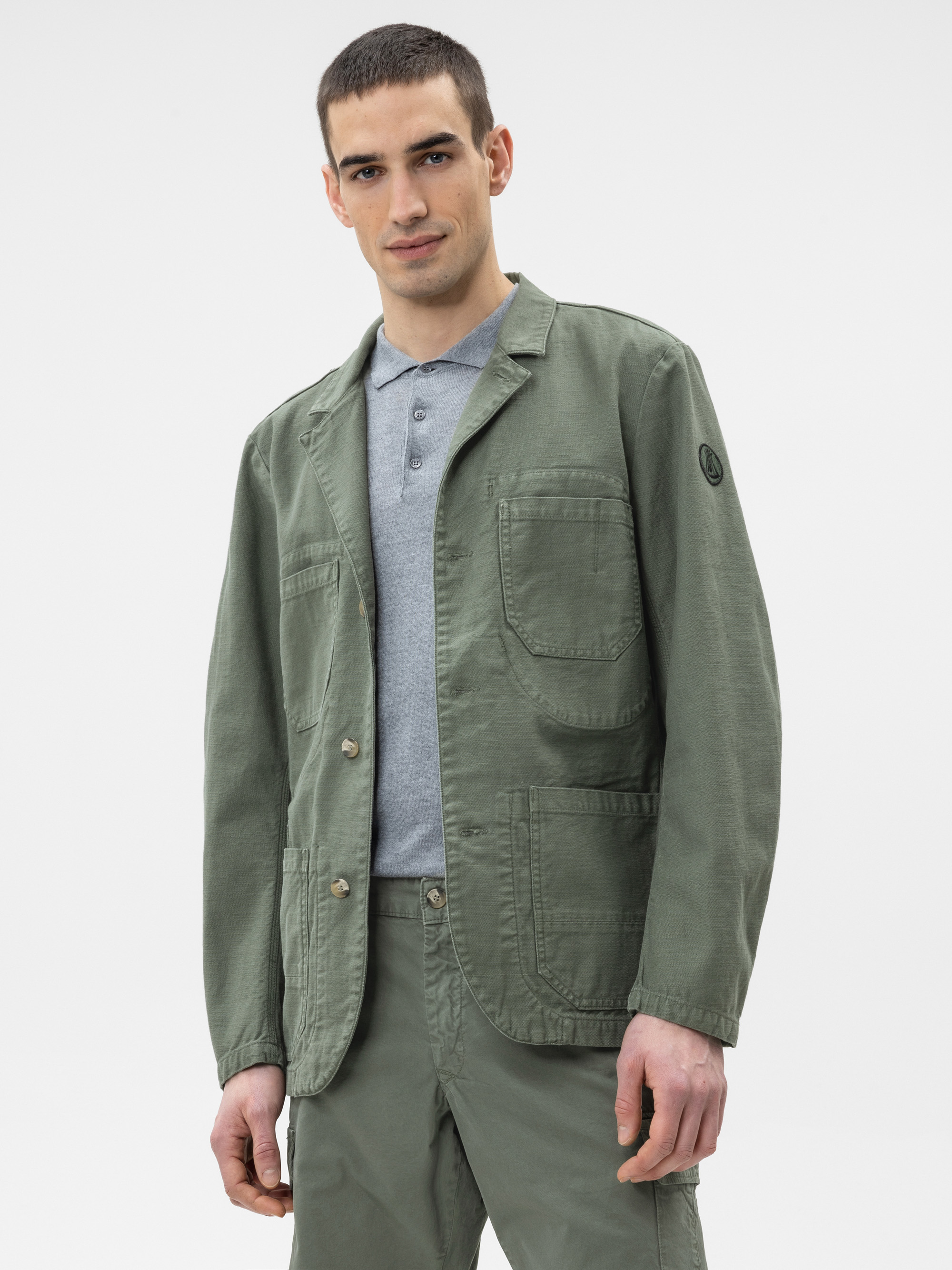 Ein Mann, der eine olivgrüne Funktionsjacke mit großen Vordertaschen über einem grauen Polohemd und einer passenden olivgrünen Hose trägt, steht vor einem schlichten weißen Hintergrund.