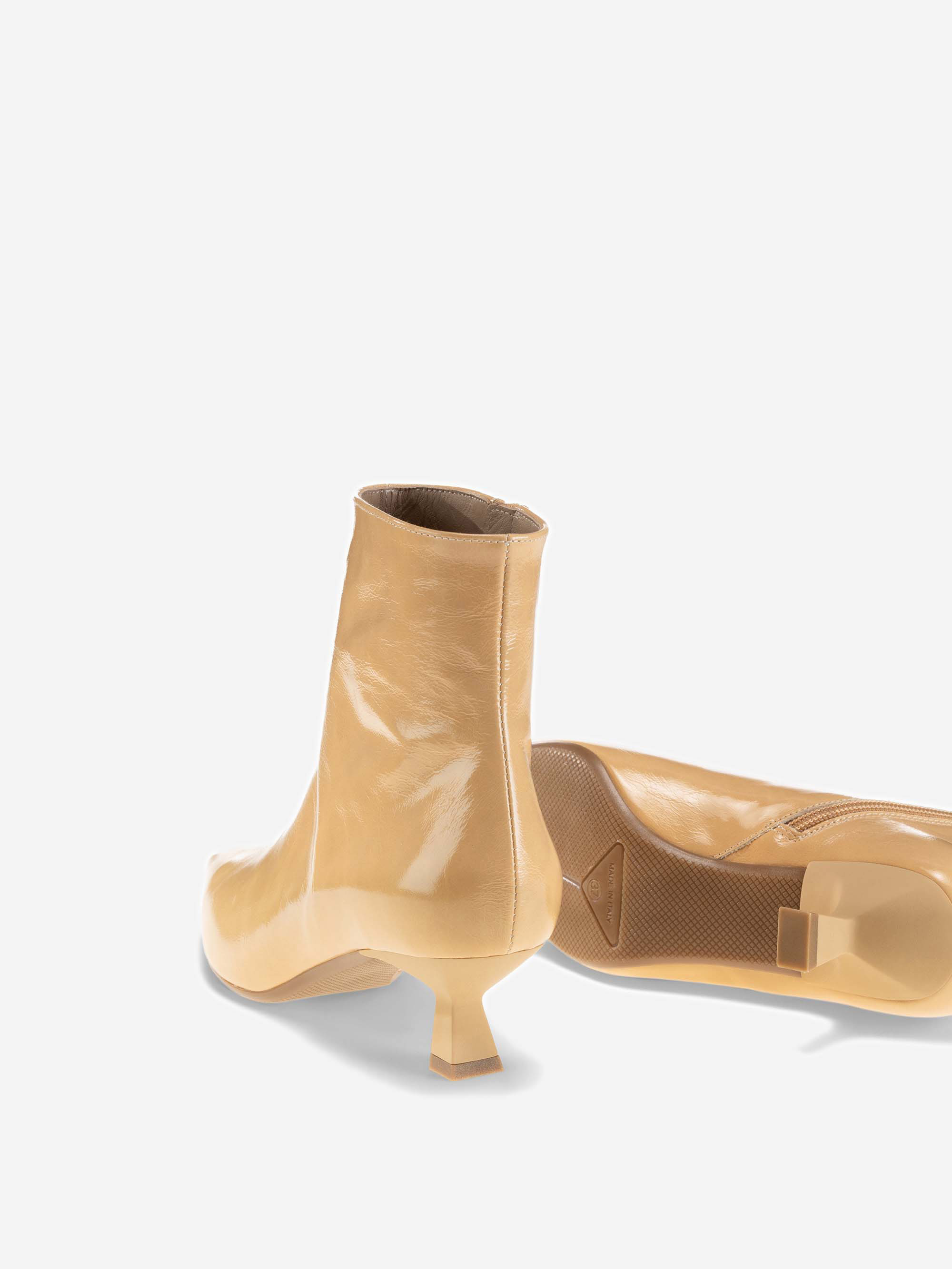 Ein Paar glänzender beiger Stiefeletten mit niedrigen, schrägen Absätzen. Ein Stiefel steht aufrecht, während der andere auf der Seite liegt und das Sohlen- und Absatzdesign vor einem schlichten weißen Hintergrund zeigt.