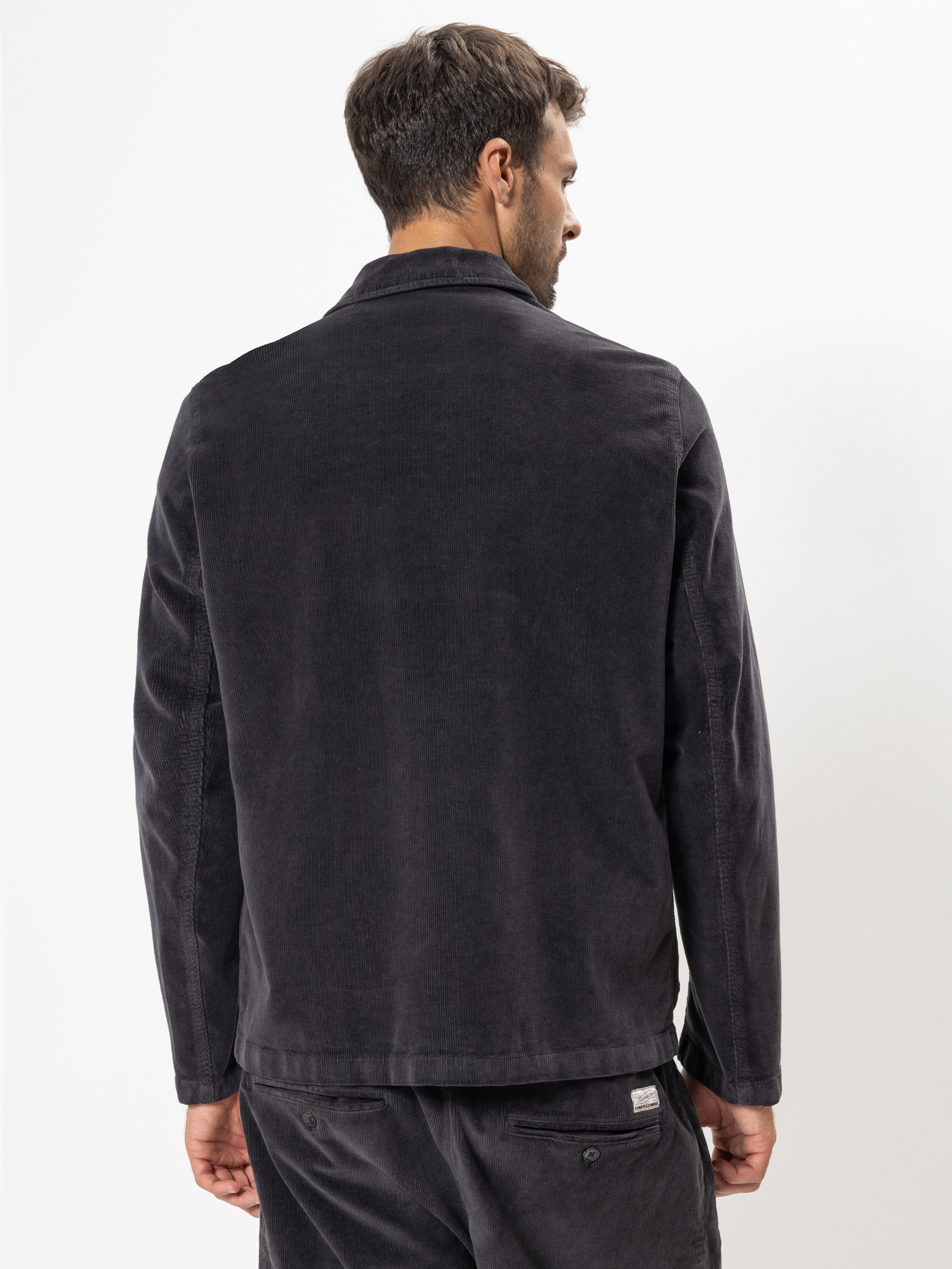 Ein Mann steht der Kamera abgewandt und trägt eine dunkle, langärmelige Cordjacke und eine passende Hose. Der Hintergrund ist einfarbig weiß.