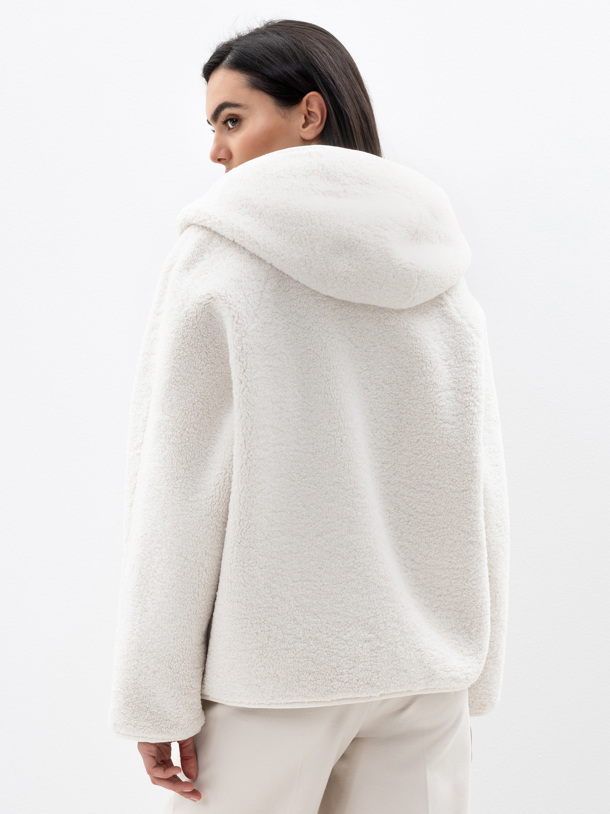 Eine Frau mit dunklem Haar trägt eine cremefarbene Sherpa-Jacke mit Kapuze und eine helle Hose. Sie steht vor einem schlichten weißen Hintergrund und schaut über ihre Schulter.