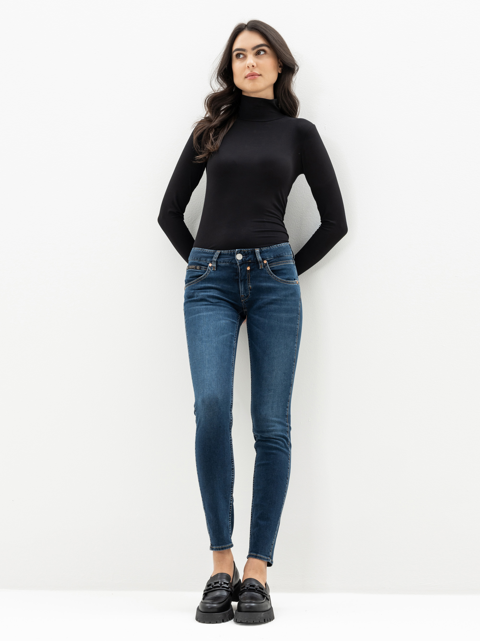 Eine Frau steht vor einem weißen Hintergrund und trägt einen schwarzen langärmeligen Rollkragenpullover, blaue Röhrenjeans und schwarze Slipper. Sie blickt leicht zur Seite und hat die Hände in den Vordertaschen.