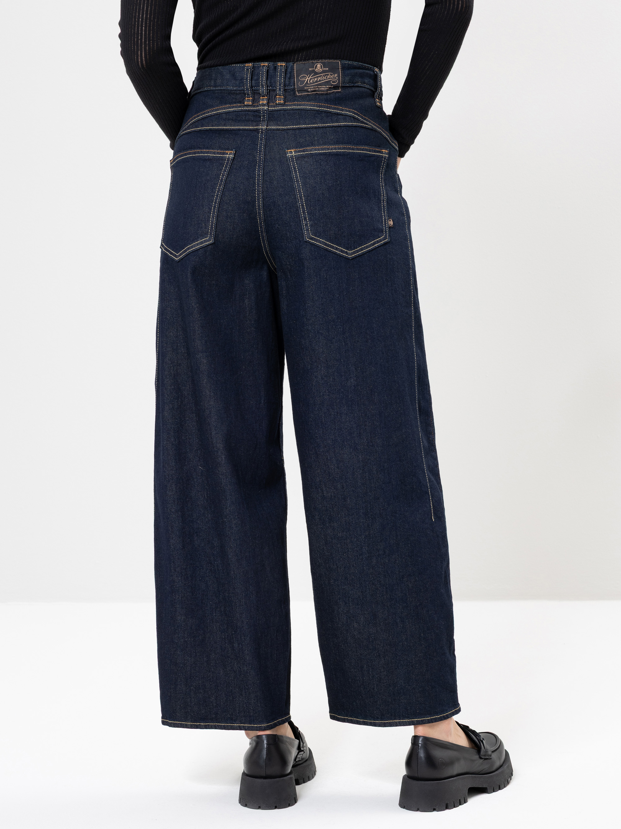 Eine Person steht mit dem Gesicht zur Wand, trägt eine dunkelblaue Jeans mit weitem Bein, ein schwarzes langärmeliges Ripp-Top und schwarze Schuhe vor einem schlichten hellen Hintergrund.