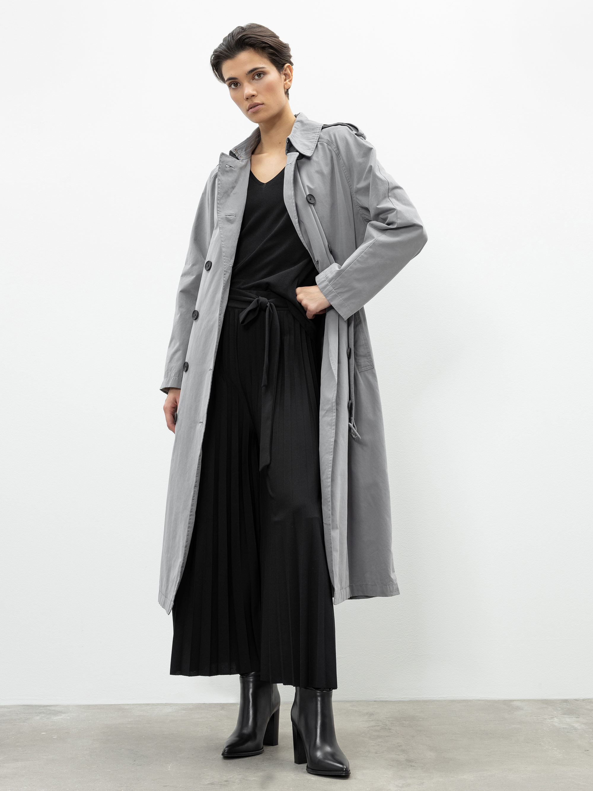 Eine Person, die einen langen grauen Trenchcoat über einem schwarzen Oberteil mit V-Ausschnitt, eine schwarze Bundfaltenhose und schwarze hochhackige Stiefel trägt, steht vor einer schlichten weißen Wand und schaut selbstbewusst in die Kamera.
