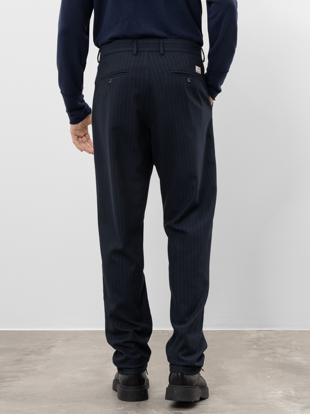 Eine Person steht mit dem Gesicht zur Kamera, trägt eine dunkle, gestreifte Anzughose, ein marineblaues langärmeliges Hemd und schwarze Schuhe vor einem schlichten hellen Hintergrund.