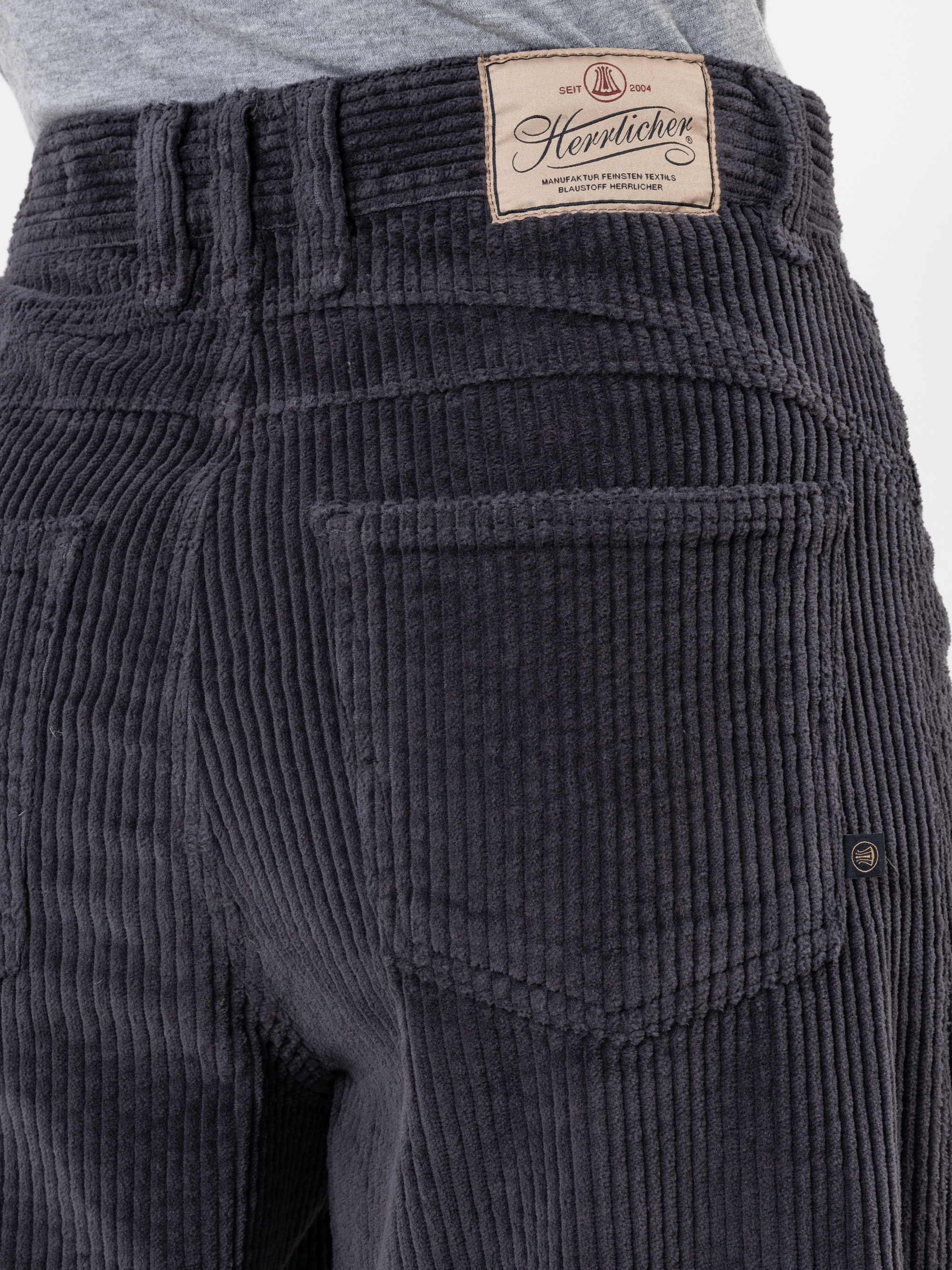 Nahaufnahme einer Person, die eine dunkelgraue Cordhose mit einer sichtbaren Gesäßtasche und einem beigen Markenetikett "Herrlicher" am Bund trägt. Die Person trägt außerdem ein hellgraues Hemd, das in die Hose gesteckt ist.