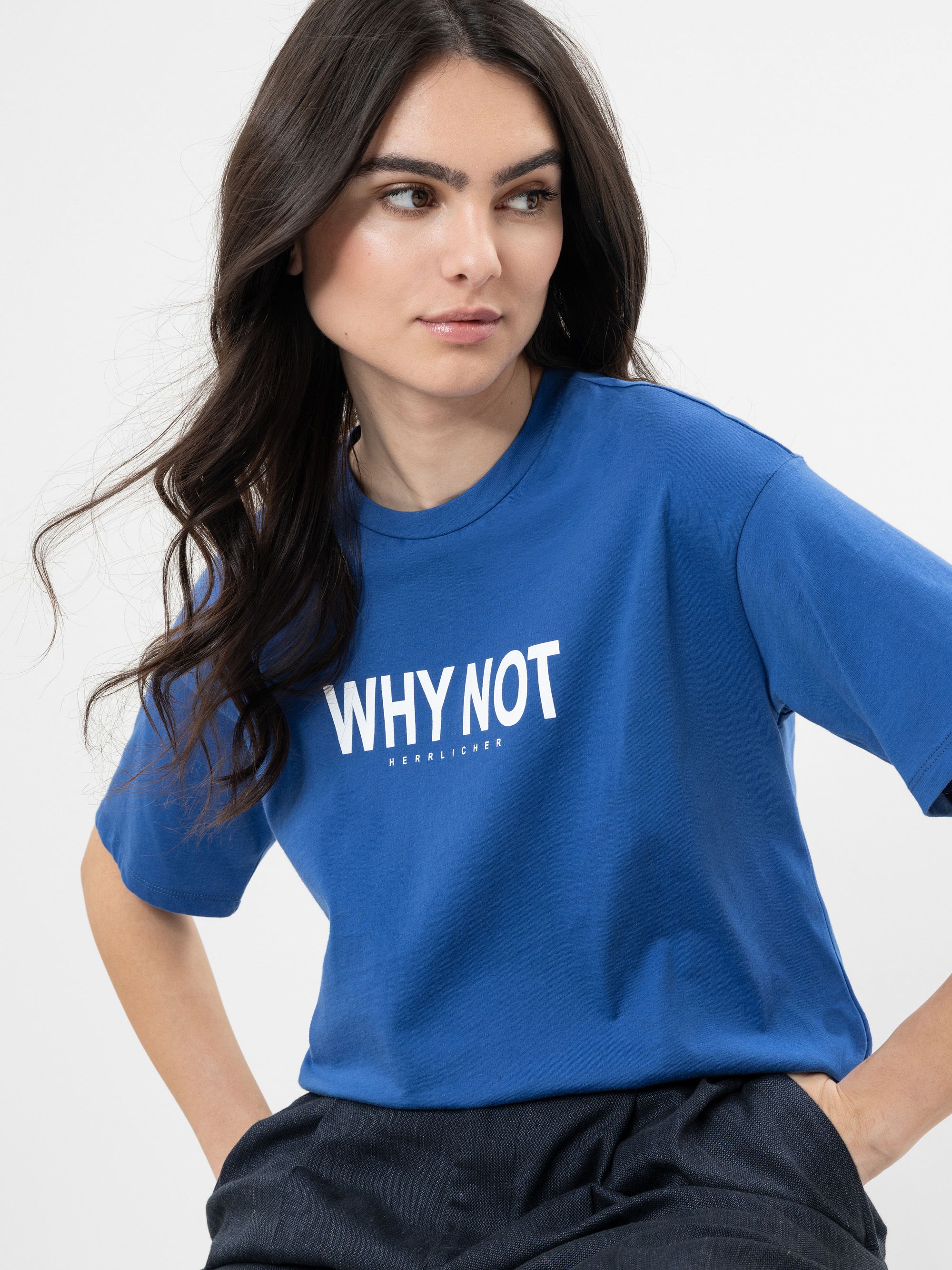 Eine Frau mit langen dunklen Haaren trägt ein blaues T-Shirt mit der Aufschrift WHY NOT in weißer Schrift auf der Vorderseite. Sie blickt mit ruhigem Blick zur Seite vor einem schlichten hellen Hintergrund.