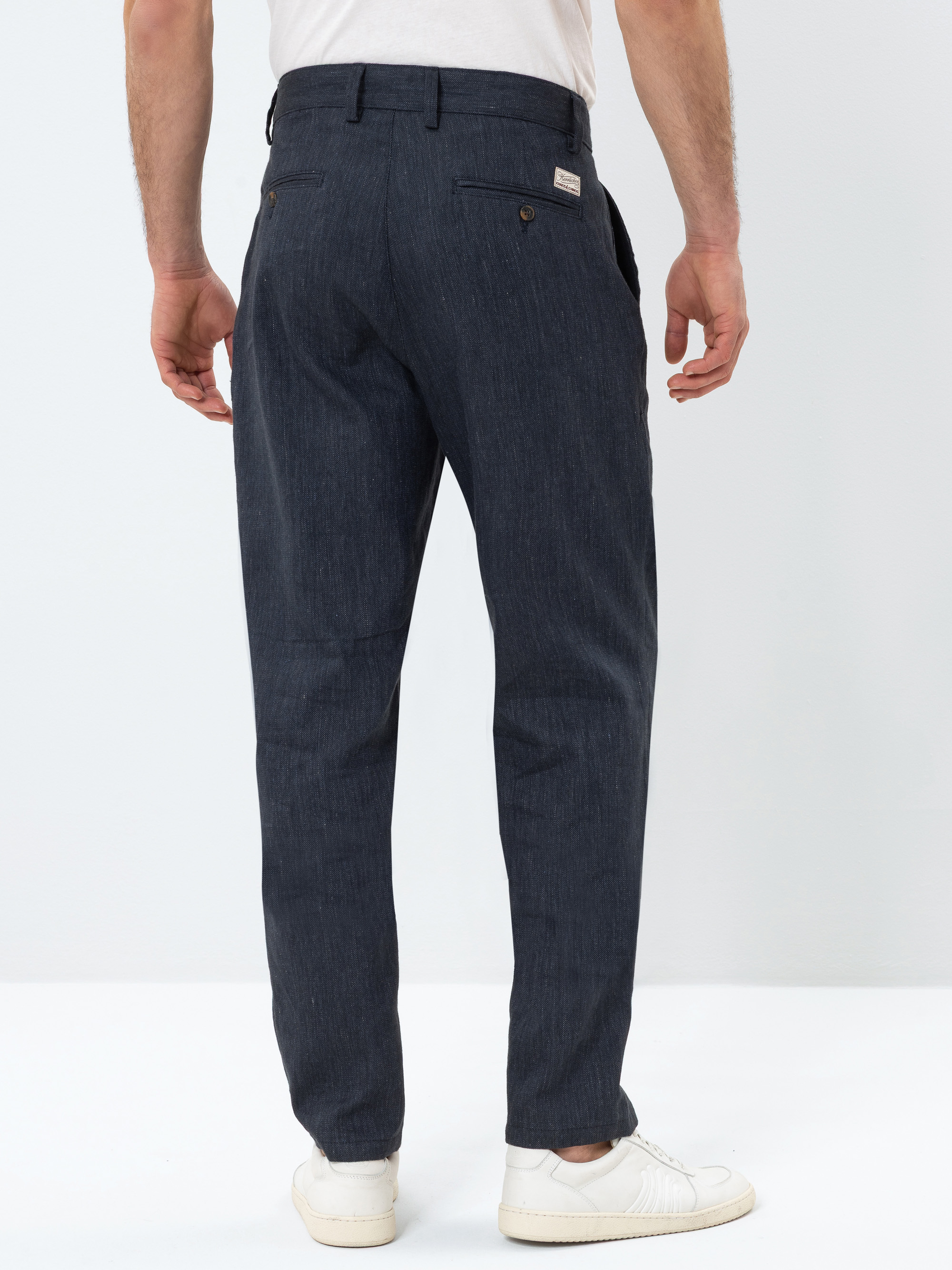 Ein Mann mit dunkelblauer Hose, weißem Hemd und weißen Turnschuhen steht mit dem Gesicht zur Wand vor einem schlichten hellen Hintergrund.