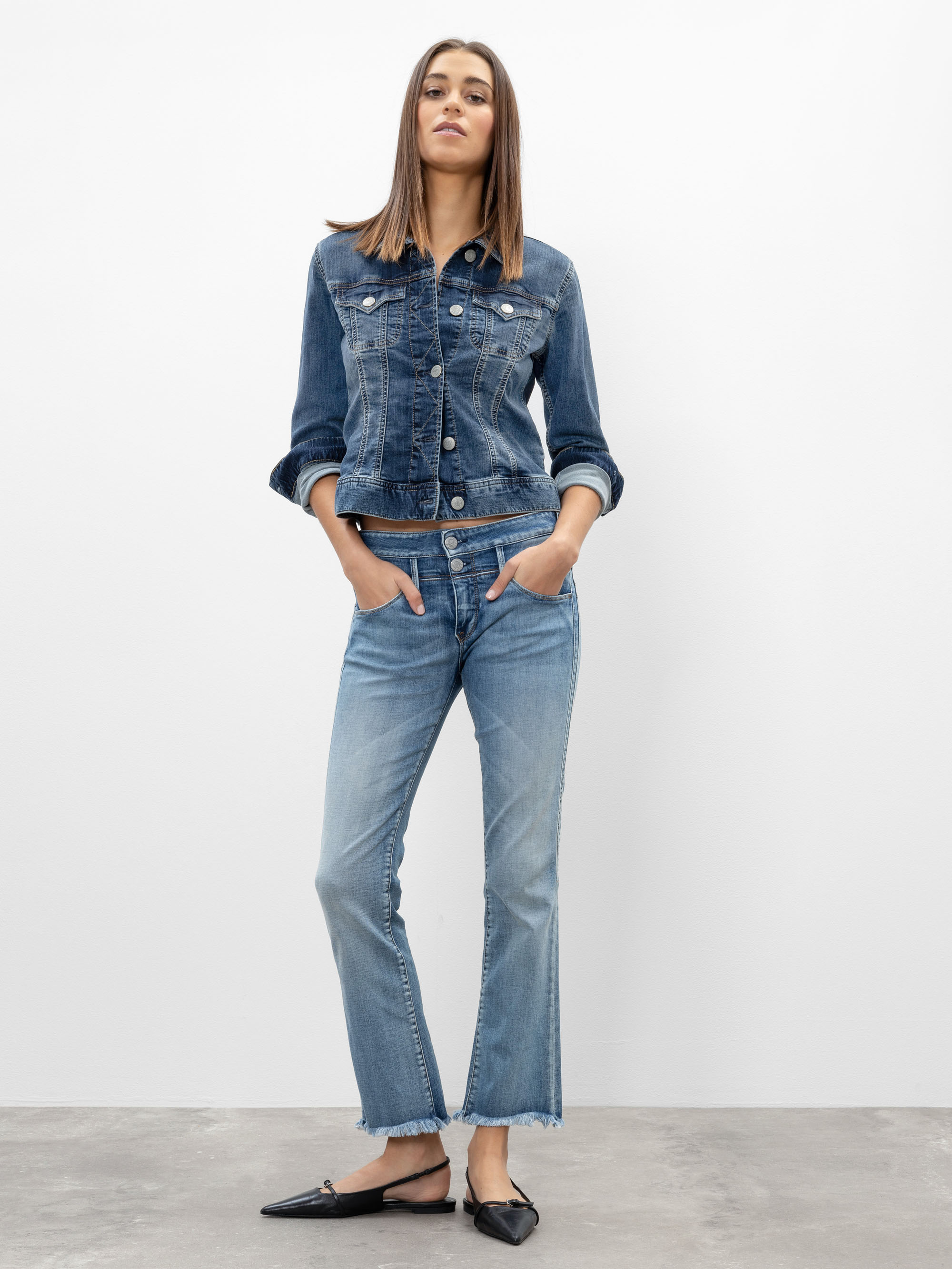 Eine Frau steht vor einem schlichten weißen Hintergrund und trägt eine blaue Jeansjacke und dazu passende blaue Jeans mit ausgefransten Säumen. Sie hat die Hände in den Taschen und trägt schwarze, spitz zulaufende Flats.