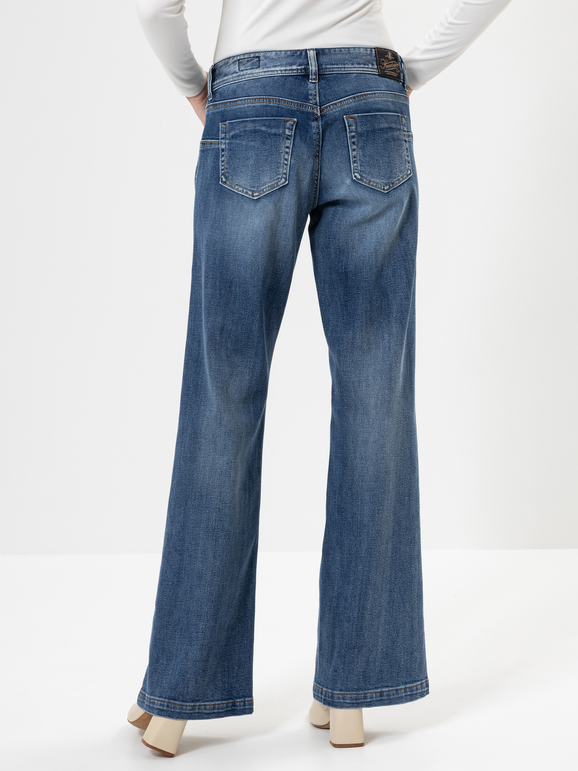 Die Person trägt eine blaue Jeans mit weitem Bein und ein weißes langärmeliges Hemd, von hinten gesehen. Die Jeans hat zwei Gesäßtaschen und ist leicht verblichen. Die Person steht vor einem weißen Hintergrund.