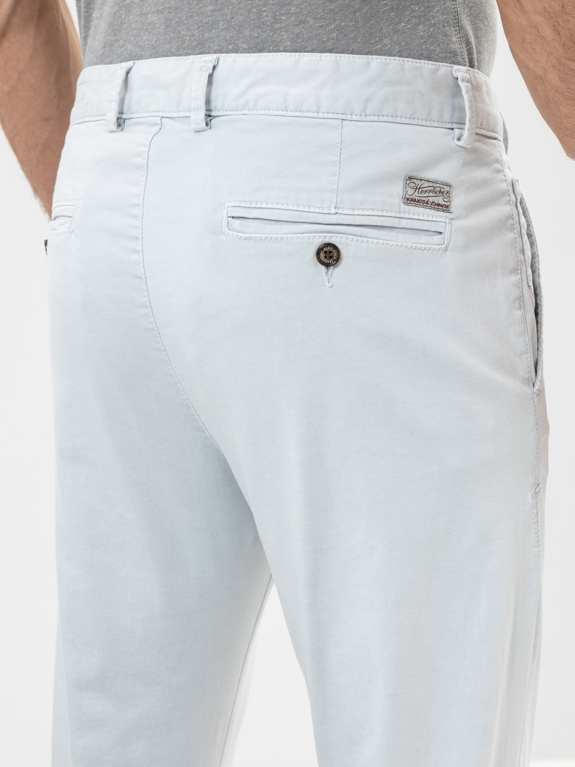 Nahaufnahme einer Person, die eine hellgraue Hose und ein graues Hemd trägt, wobei die Gesäßtasche mit einem Knopf und einem kleinen Markenetikett über der Tasche zu sehen ist.