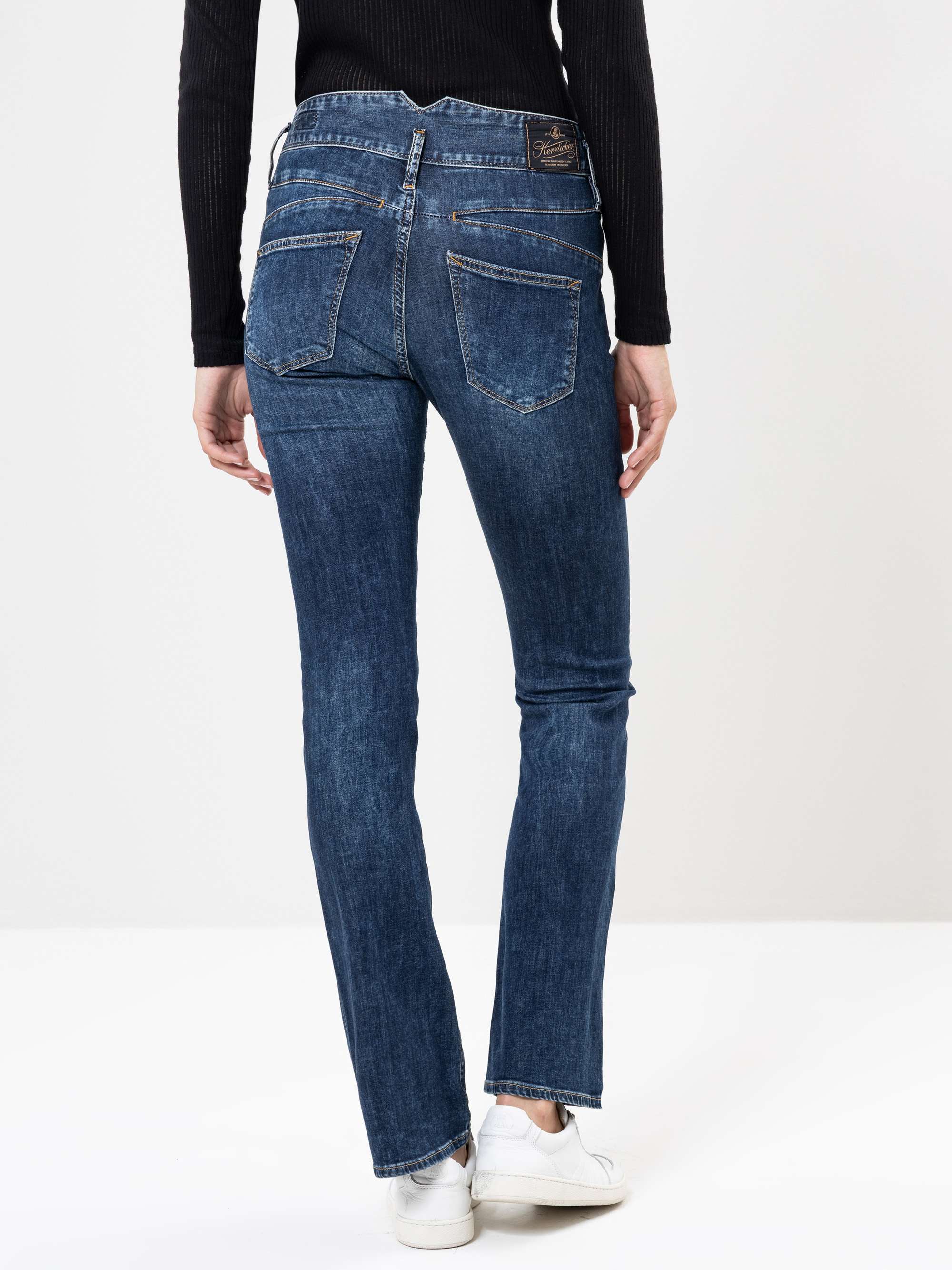 Eine Person, die einen schwarzen Pullover und blaue Jeans trägt, steht mit dem Rücken zur Kamera. Sie trägt außerdem weiße Turnschuhe. Der Hintergrund ist einfarbig und hell.