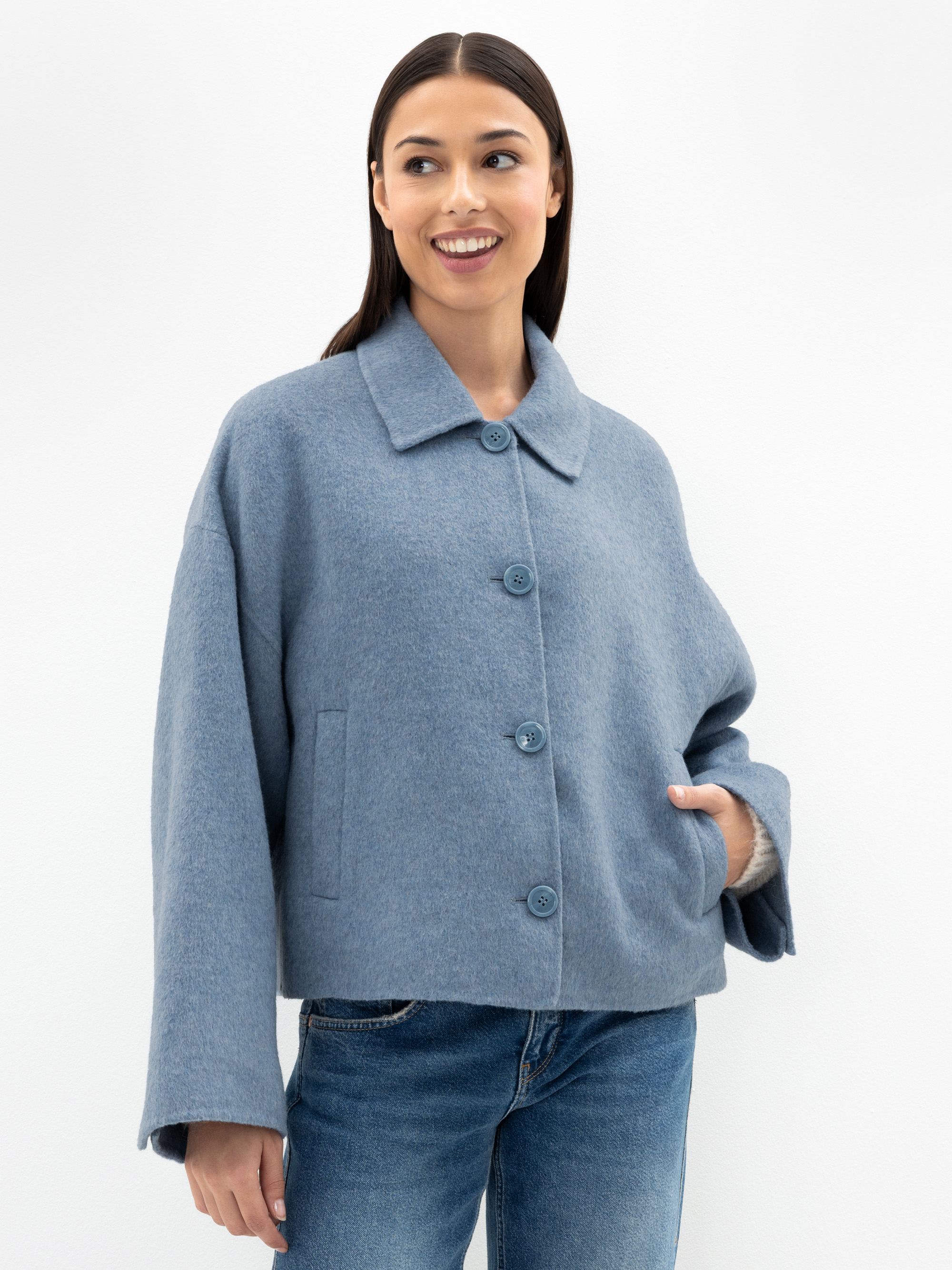 Eine Frau mit glattem dunklem Haar trägt einen hellblauen Wollmantel mit Knöpfen und blaue Jeans und steht lächelnd vor einem schlichten weißen Hintergrund.