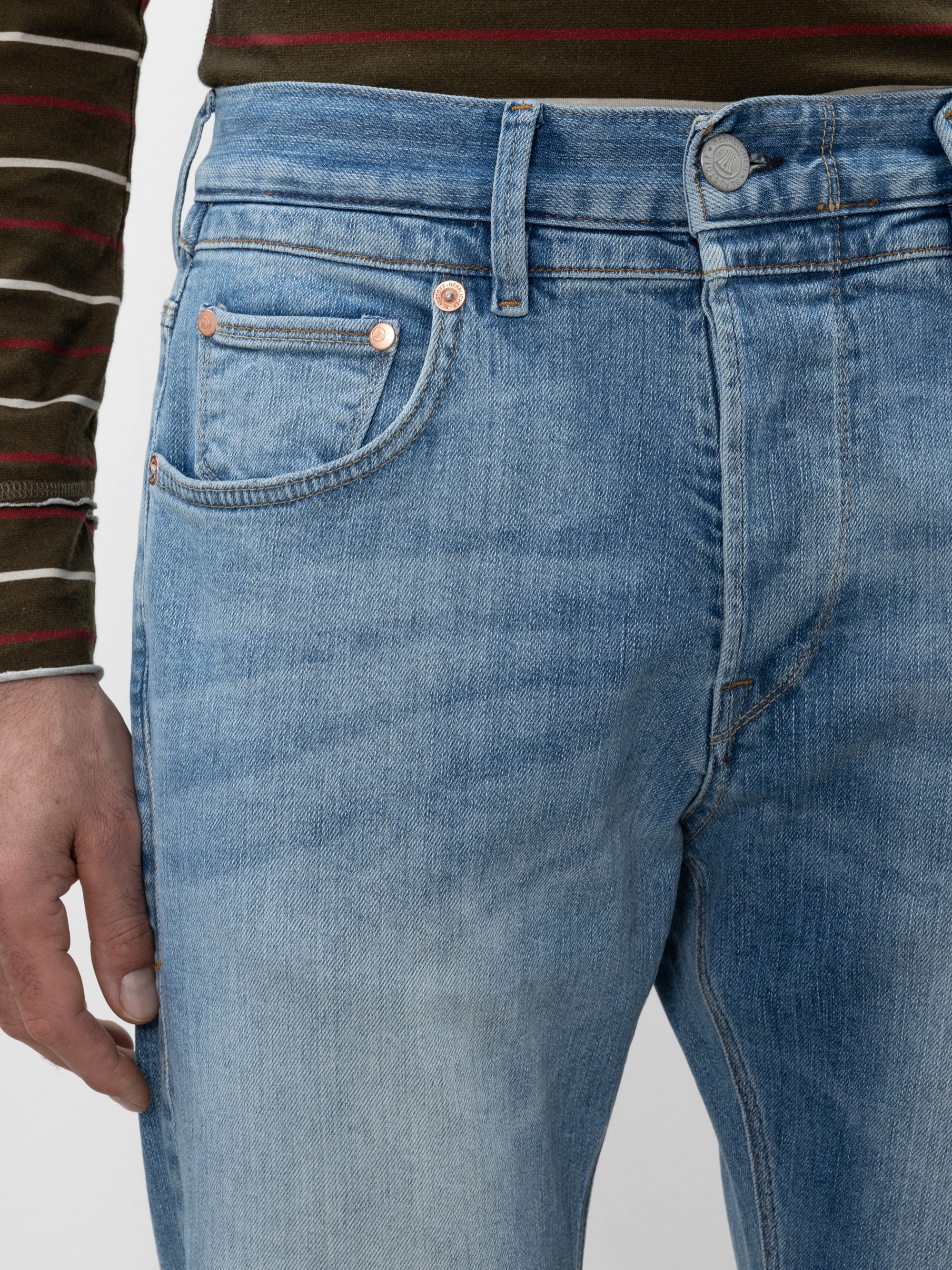 Eine Person trägt eine hellblaue Jeans und ein grün und braun gestreiftes langärmeliges Hemd, die linke Hand liegt entspannt an der Seite. Das Bild konzentriert sich auf den Bund der Jeans und den Bereich der Oberschenkel.