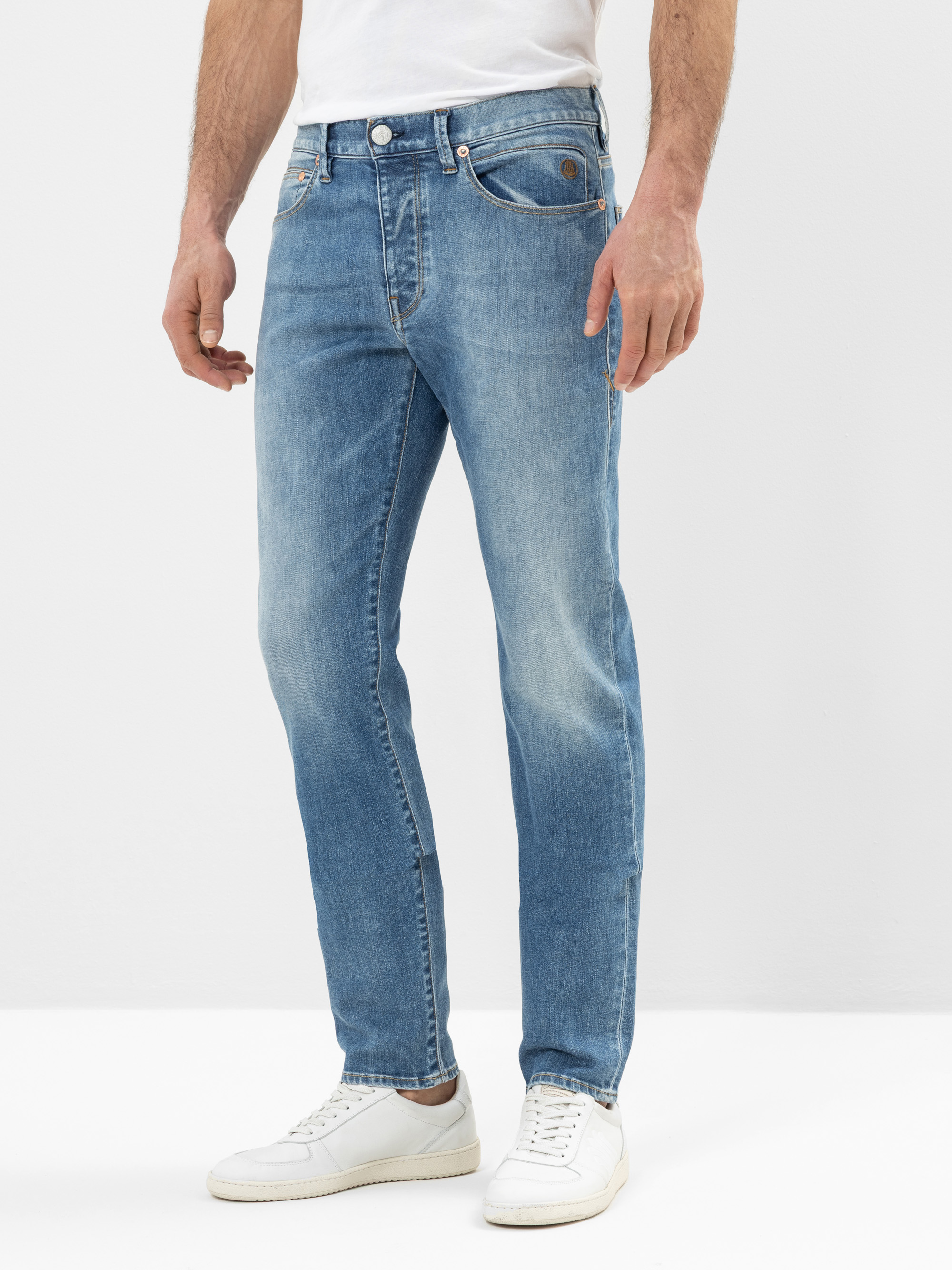 Ein Mann steht vor einem schlichten Hintergrund, trägt hellblaue Jeans mit geradem Bein und weiße Turnschuhe, die Hände liegen entspannt an den Seiten.
