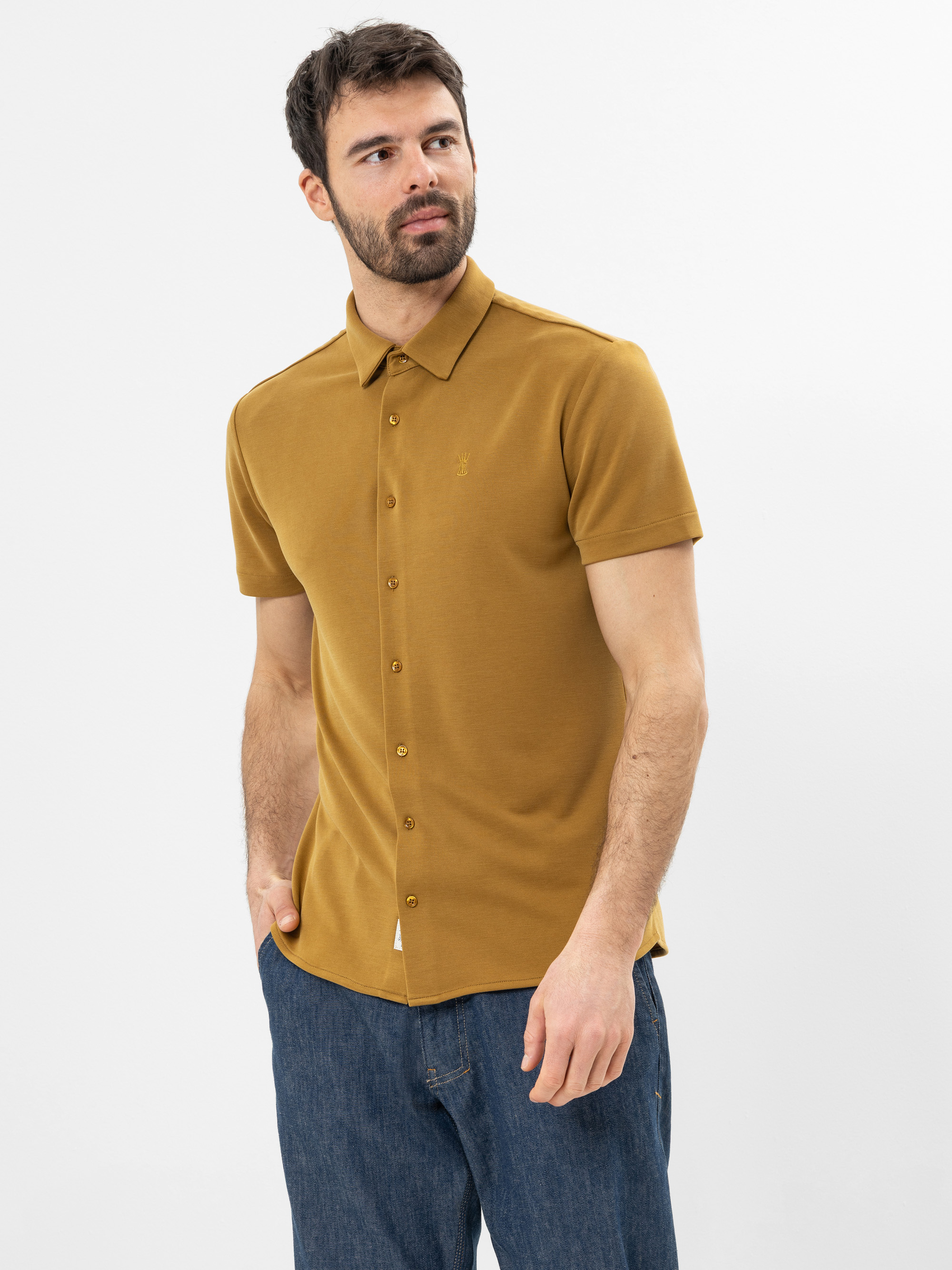 Ein Mann mit kurzen dunklen Haaren und einem Bart trägt ein senfgelbes, kurzärmeliges Button-up-Hemd und blaue Jeans. Er steht vor einem schlichten weißen Hintergrund und hat eine Hand in der Tasche.