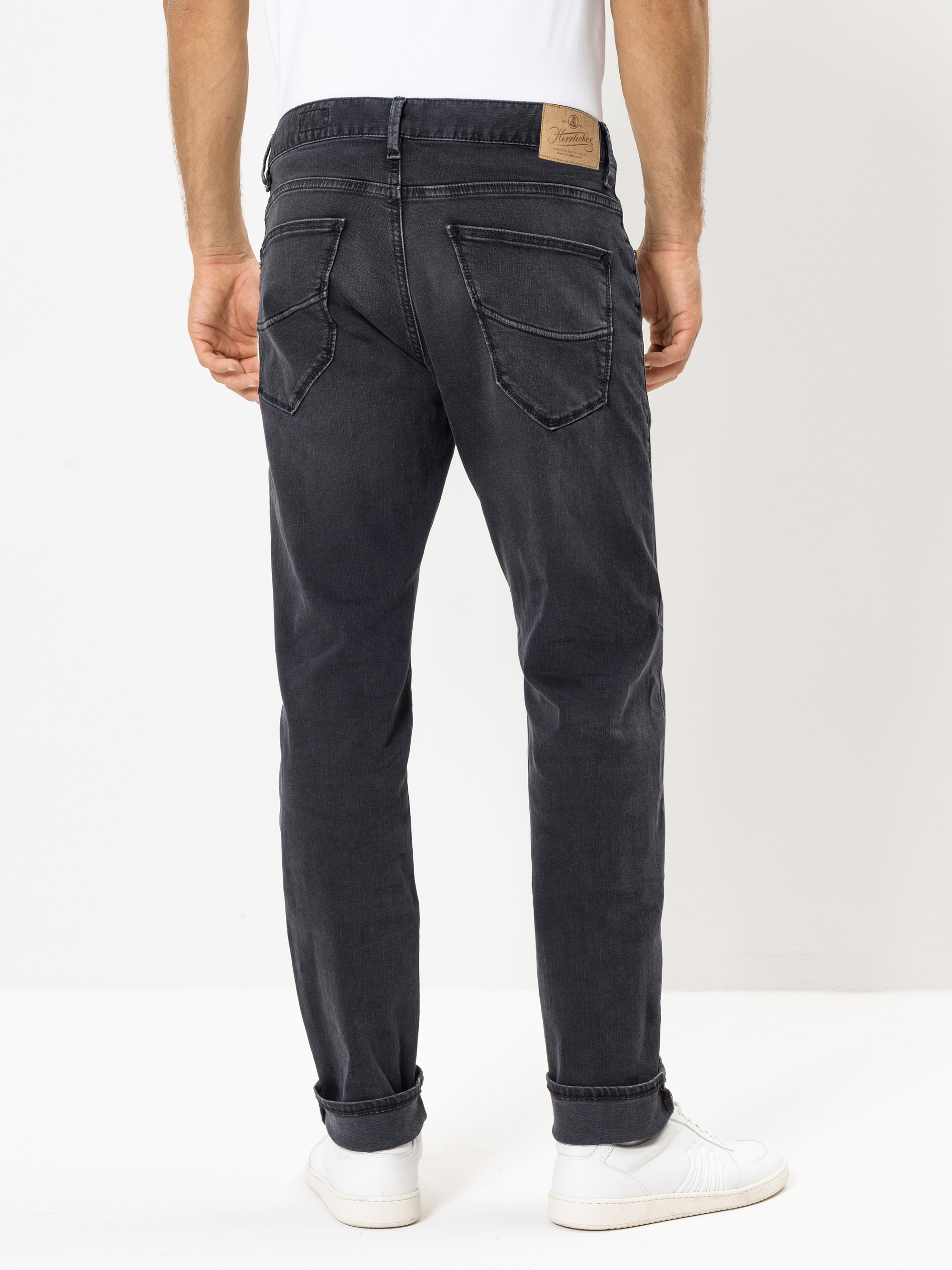 Eine Person, die schwarze Jeans, ein weißes T-Shirt und weiße Turnschuhe trägt, steht mit dem Rücken zur Kamera vor einem schlichten weißen Hintergrund.