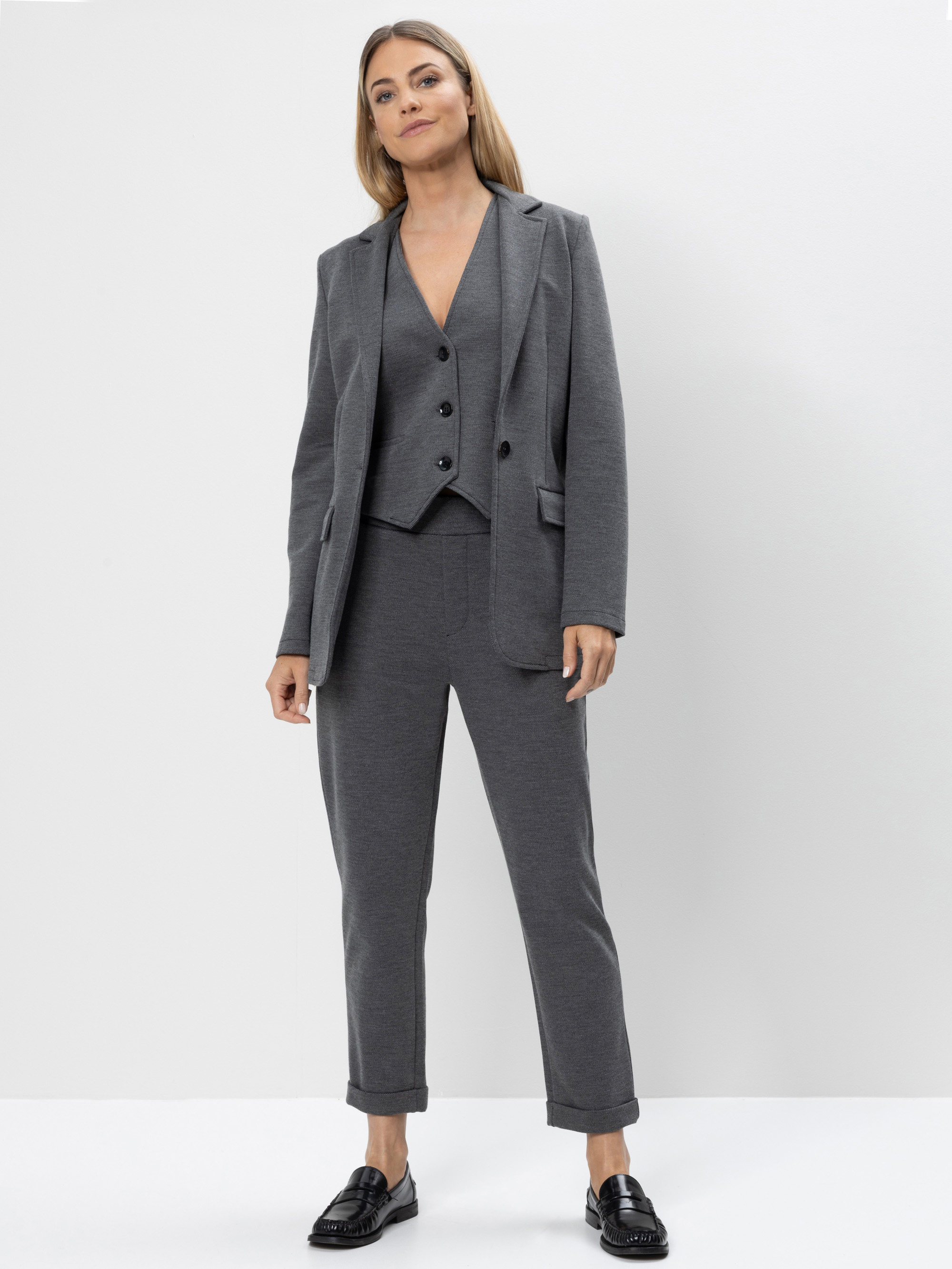 Eine Frau in einem grauen Anzug mit Blazer, passender Weste und Hose steht selbstbewusst vor einem schlichten weißen Hintergrund. Sie hat glattes hellbraunes Haar und trägt schwarze Halbschuhe.