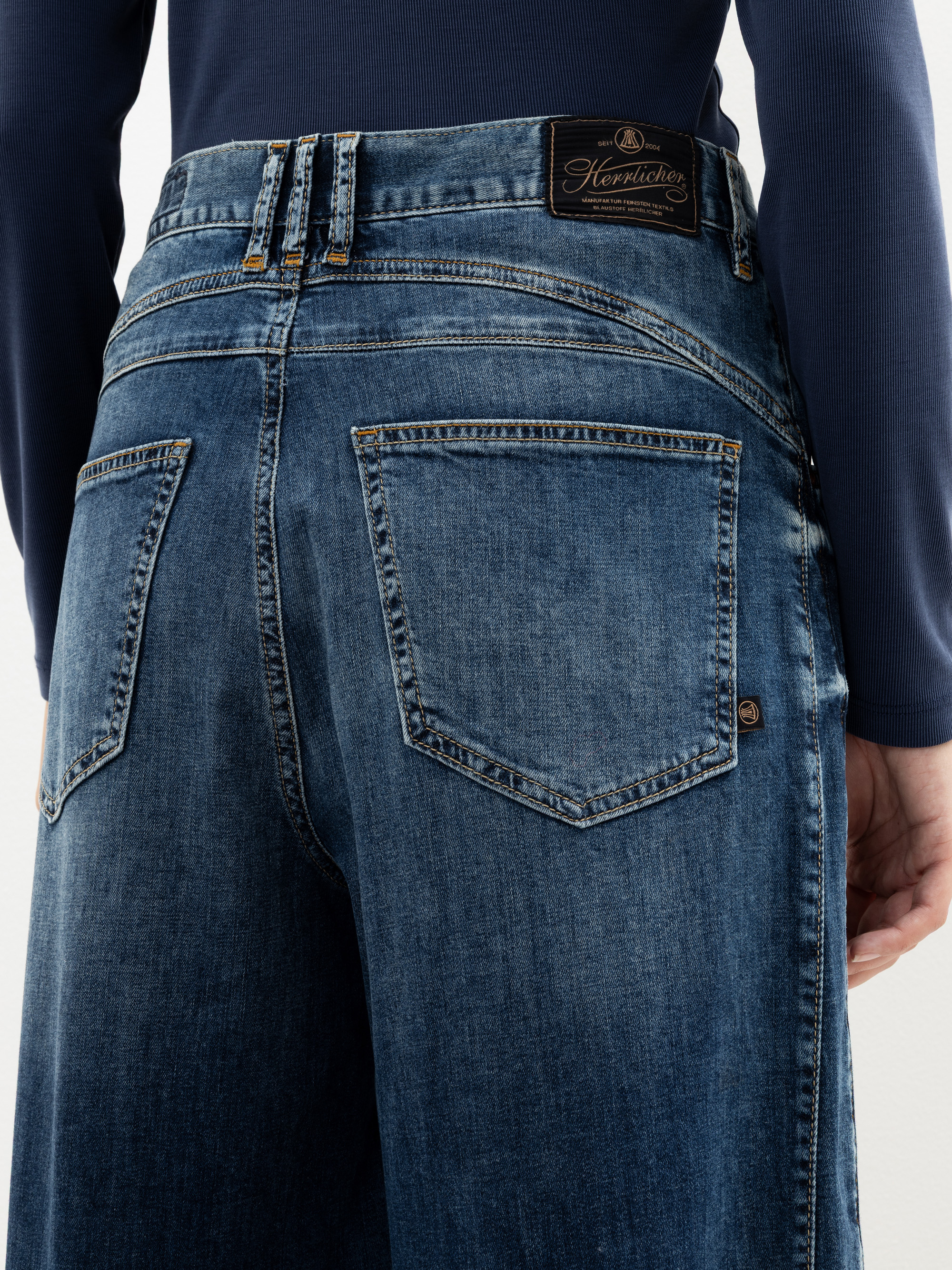 Eine Person, die eine blaue Jeans mit weitem Bein und ein marineblaues langärmeliges Oberteil trägt, wird von hinten gezeigt. Die Jeans hat große Gesäßtaschen, sichtbare Nähte und einen Markenaufnäher aus Leder am Bund.