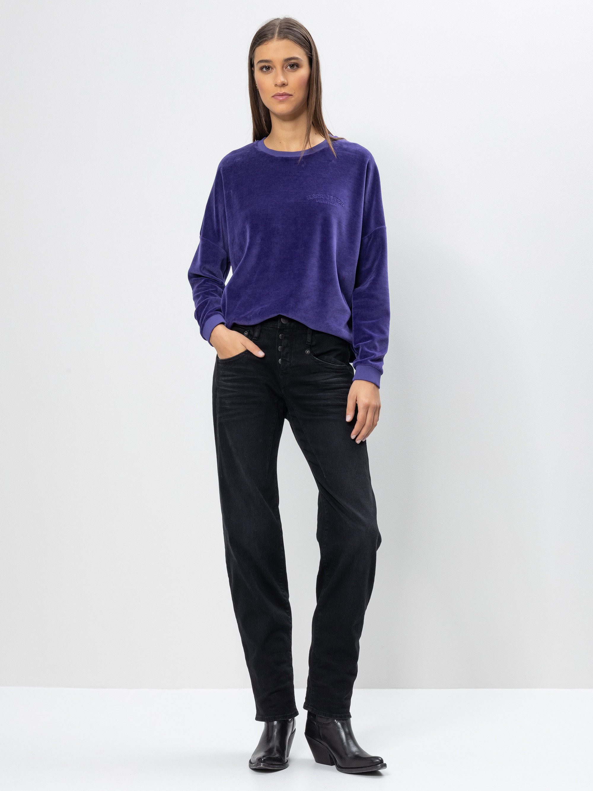 Eine Frau steht vor einem schlichten weißen Hintergrund und trägt ein locker sitzendes, langärmeliges lilafarbenes Oberteil, schwarze Jeans mit hoher Taille und schwarze Ankle Boots. Sie hat glattes, braunes Haar und einen neutralen Gesichtsausdruck.